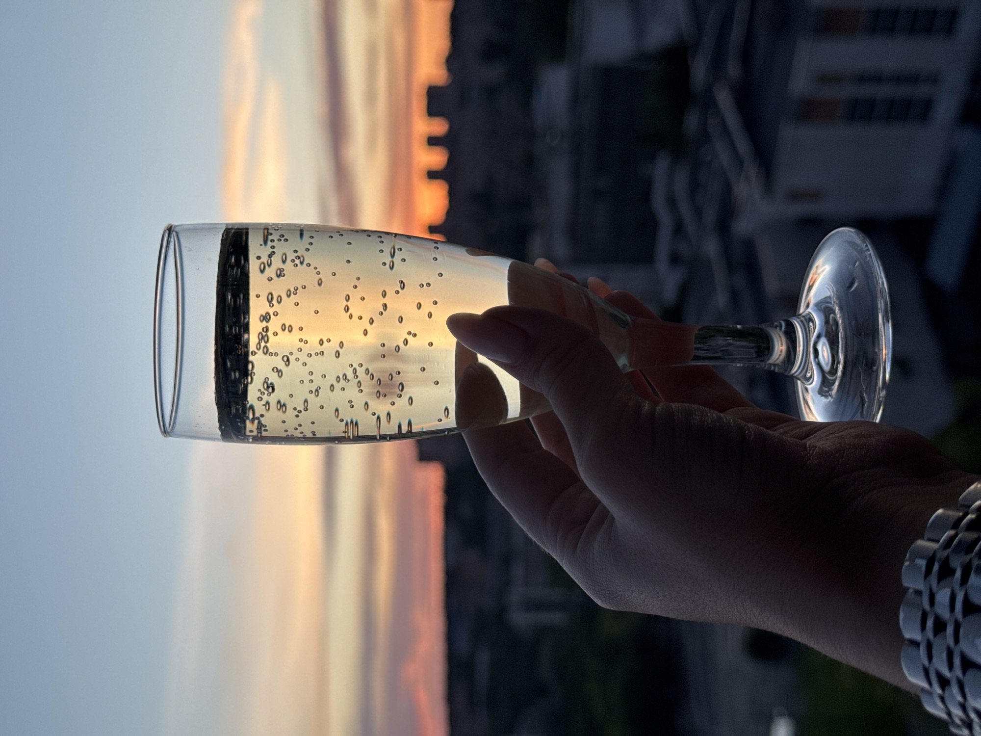 A hand holds a champagne flute filled with sparkling liquid against a sunset backdrop. Bubbles are visible within the drink. The background appears to show a blurred cityscape with some structures illuminated by the setting sun. A metal watch bracelet is visible on the wrist of the hand holding the glass. It is not possible to confirm if this image was taken in Izhevsk, Russia based on the provided information.