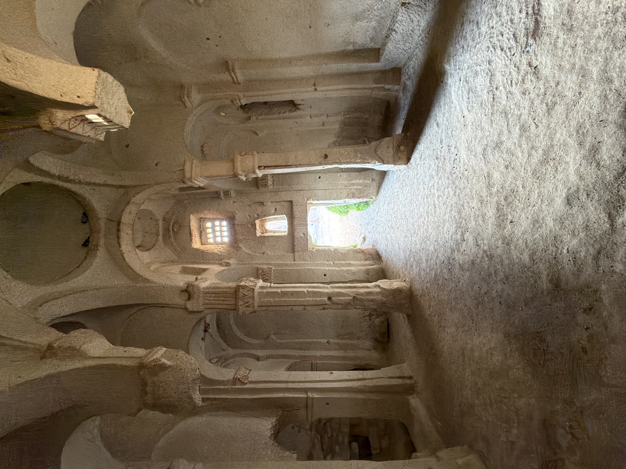 The image displays the interior of a rock-cut structure, likely a cave church or dwelling, characteristic of Ürgüp, Turkey. Carved entirely from light-colored, rough-hewn stone, the space features a series of robust, cylindrical pillars with elaborately carved capitals supporting arches and a vaulted ceiling. Some capitals are adorned with distinct zigzag patterns. The uneven surfaces of the walls and ceiling emphasize the hand-carved nature of the architecture.
Natural light illuminates the chamber through several small, square-gridded windows, creating strong contrasts of light and shadow that highlight the stone's texture. A larger, brightly lit opening at what appears to be the far end leads outdoors, revealing vibrant green foliage. This opening suggests an exit or a connection to another part of the rock complex. The floor area, visible on the right side of the image, is a relatively flat, textured rock surface. No individuals or specific activities are present, giving the scene a timeless and serene quality. The image effectively conveys the unique craftsmanship and historical ambiance of the Cappadocian region. No visible text can be discerned.