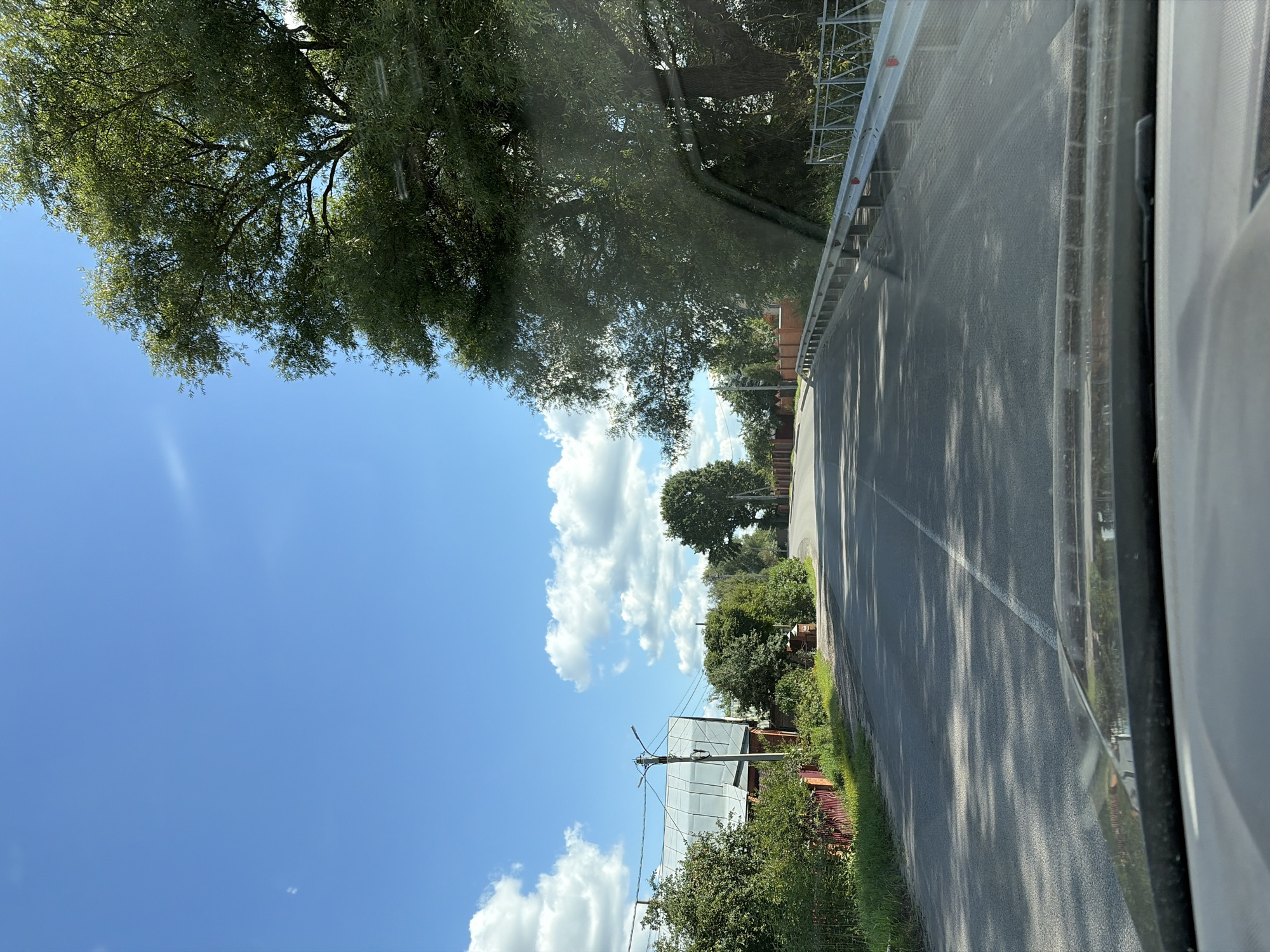 The image shows a road in the city of Novoye Podvyaznovo, Russia, on a sunny day with a blue sky and scattered clouds. The road is paved and has a white line marking the center. On the left side of the road, there are houses, trees, and a red fence. On the right side of the road, there is a metal guardrail and more trees. The image appears to be taken from inside a vehicle, as the dashboard is visible in the foreground. The overall scene depicts a rural or suburban setting in Russia.