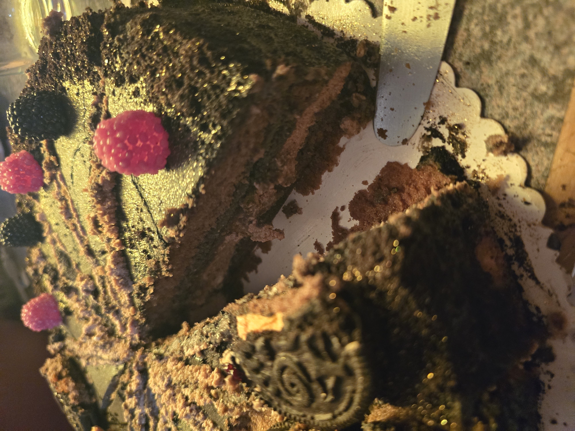 The image shows a partially cut chocolate cake. It is decorated with raspberries and what appear to be blackberries or chocolate decorations resembling blackberries. A piece of the cake has been removed, revealing the layers. A silver serving utensil is visible near the cut edge of the cake. The cake sits on a white, scalloped-edged cake board. There is no indication of the location being Zirndorf, Germany. The image focuses on the cake; there are no people or specific environmental clues.