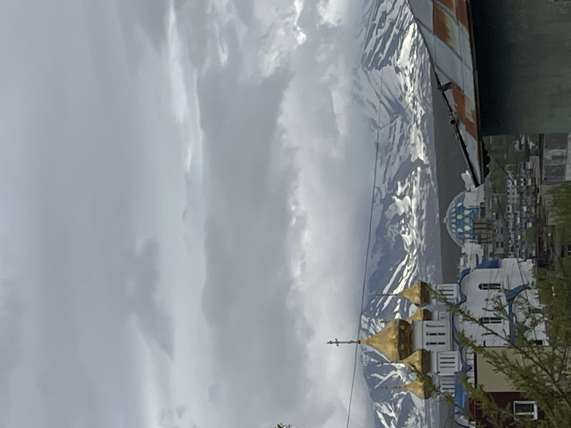 The image captures an overcast day in Petropavlovsk-Kamchatsky, Russia, with a view of the cityscape against a backdrop of snow-capped mountains. In the foreground, a white and blue Orthodox church with multiple golden domes is prominently featured. To the right, a weathered, rusty metal roof of a building juts into the frame. In the middle ground, a distinctive blue-domed building is visible, along with other urban structures. The background is dominated by the rugged, snow-covered peaks of the volcanoes surrounding the city. The sky is heavy with grey clouds, suggesting a cool or rainy climate. The composition offers a blend of religious architecture, urban development, and the imposing natural landscape of the Kamchatka Peninsula.
