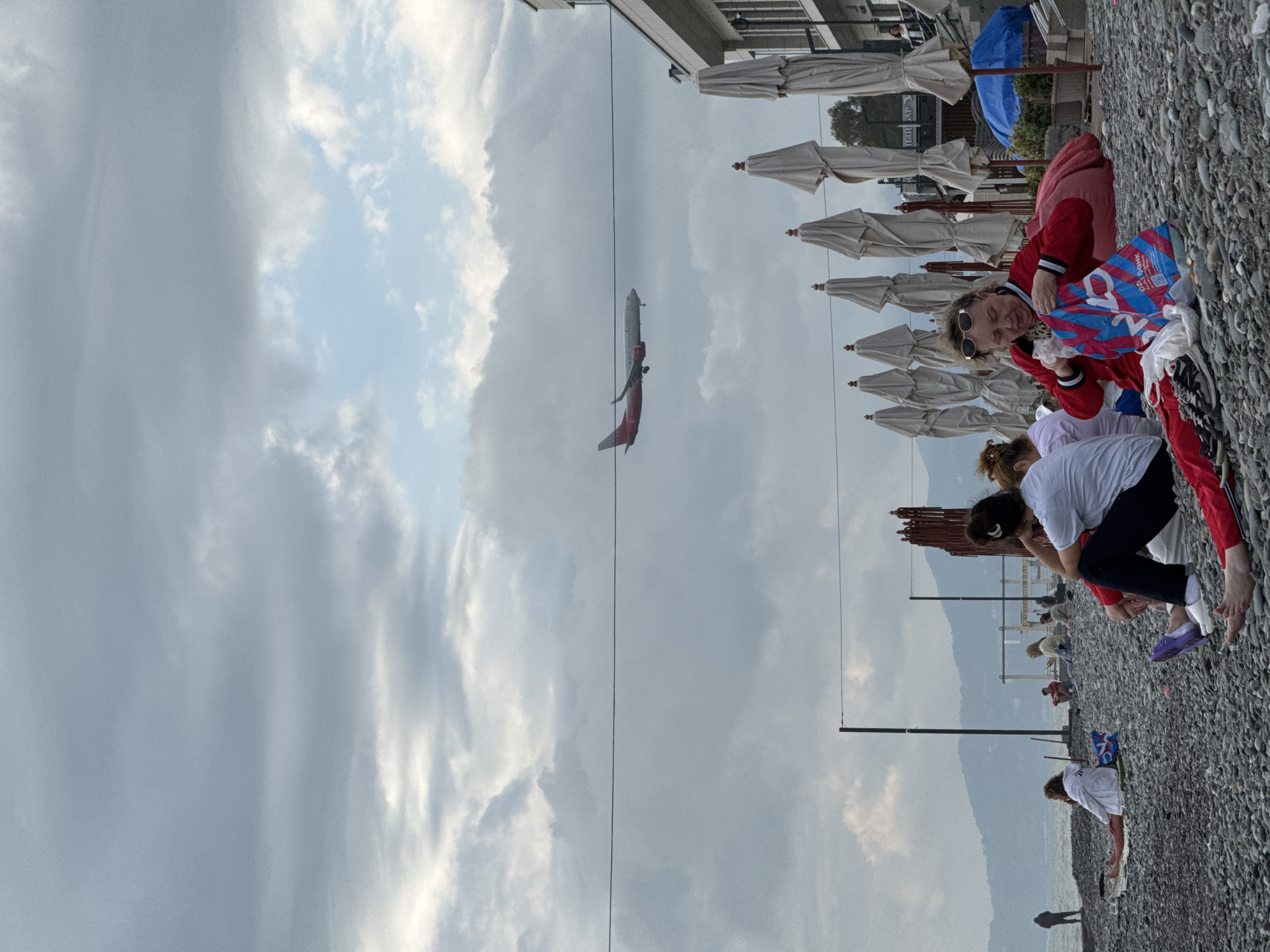 On a pebbled beach in Sochi, Russia, several people are present, near white umbrellas. In the air, an airplane is visible. One woman is near a blue bag that says "OZON." Mountains are in the distance. The sky is cloudy. Overhead power lines are visible.