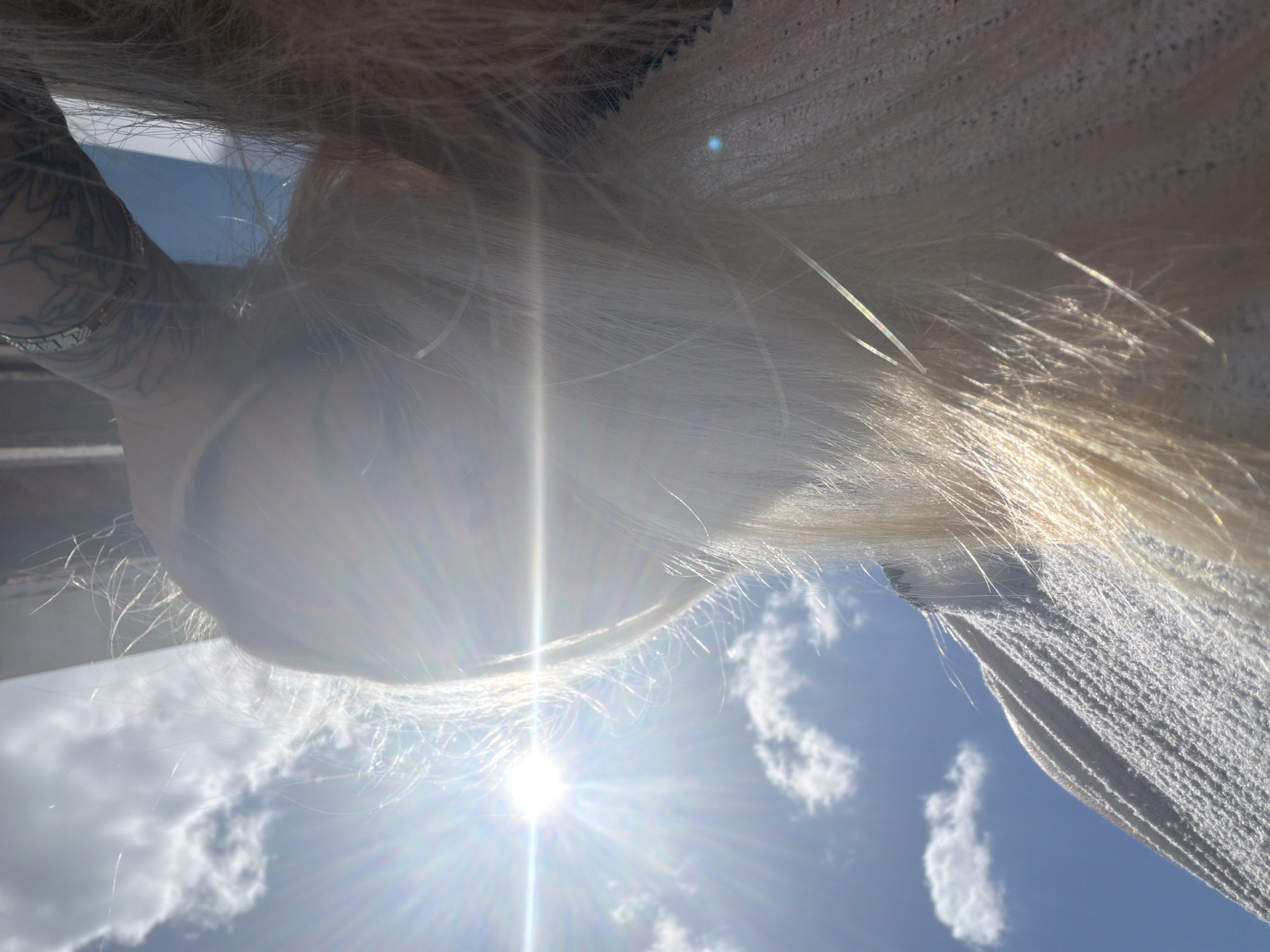 The image depicts a close-up, low-angle view of a person with blonde hair in Sarapul, Russia. The sun is shining brightly, creating a lens flare across the image. The person appears to be wearing a light-colored, textured sweater. A tattooed arm and shoulder are visible in the upper right corner. The background consists of a bright blue sky with scattered white clouds and a dark, possibly metallic, structure above. The overall impression is one of a candid, outdoor moment with strong natural light.