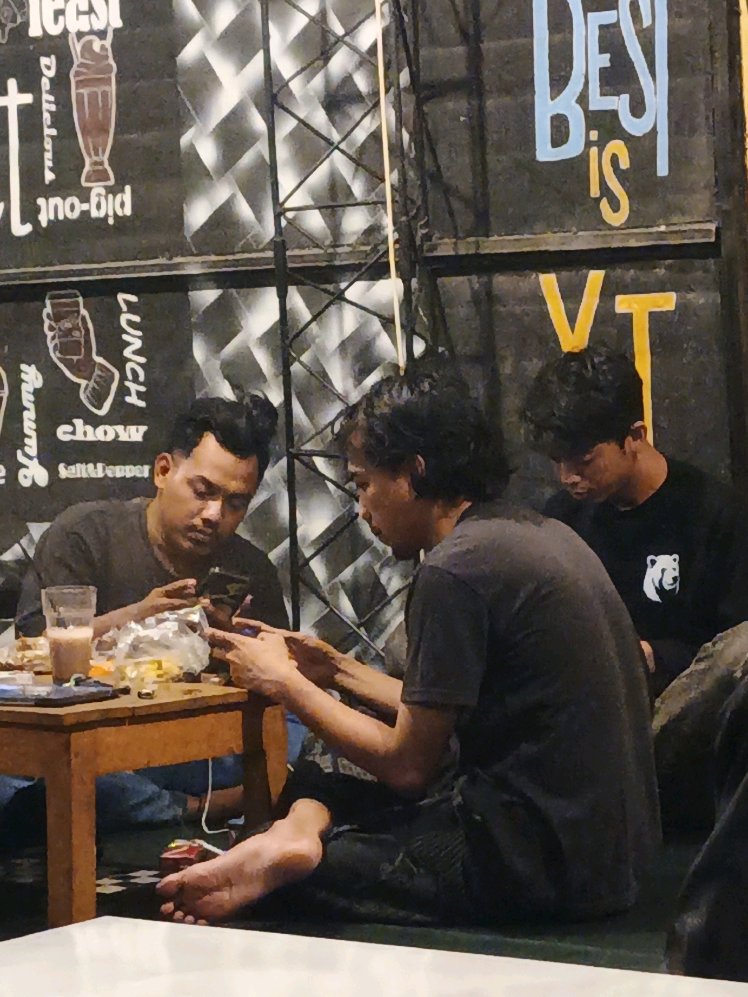 The image depicts a dimly lit indoor setting, possibly a cafe or restaurant, in Ketegan, Indonesia. Three individuals are visible, all appearing to be young adult males. The foreground shows a small, wooden table laden with various items including a glass of drink, food packaging, and cell phones. One individual in the center foreground is seated on the floor, barefoot, intensely focused on his phone, with his feet positioned on the floor. Another individual to his left is also seated, holding a phone, and seemingly engaged with it. A third individual is partially visible on the right, wearing a black t-shirt with a white bear logo. The background features a dark wall with white lettering and graphics, some of which are partially obscured. The overall atmosphere suggests a casual gathering where the individuals are preoccupied with their mobile devices.