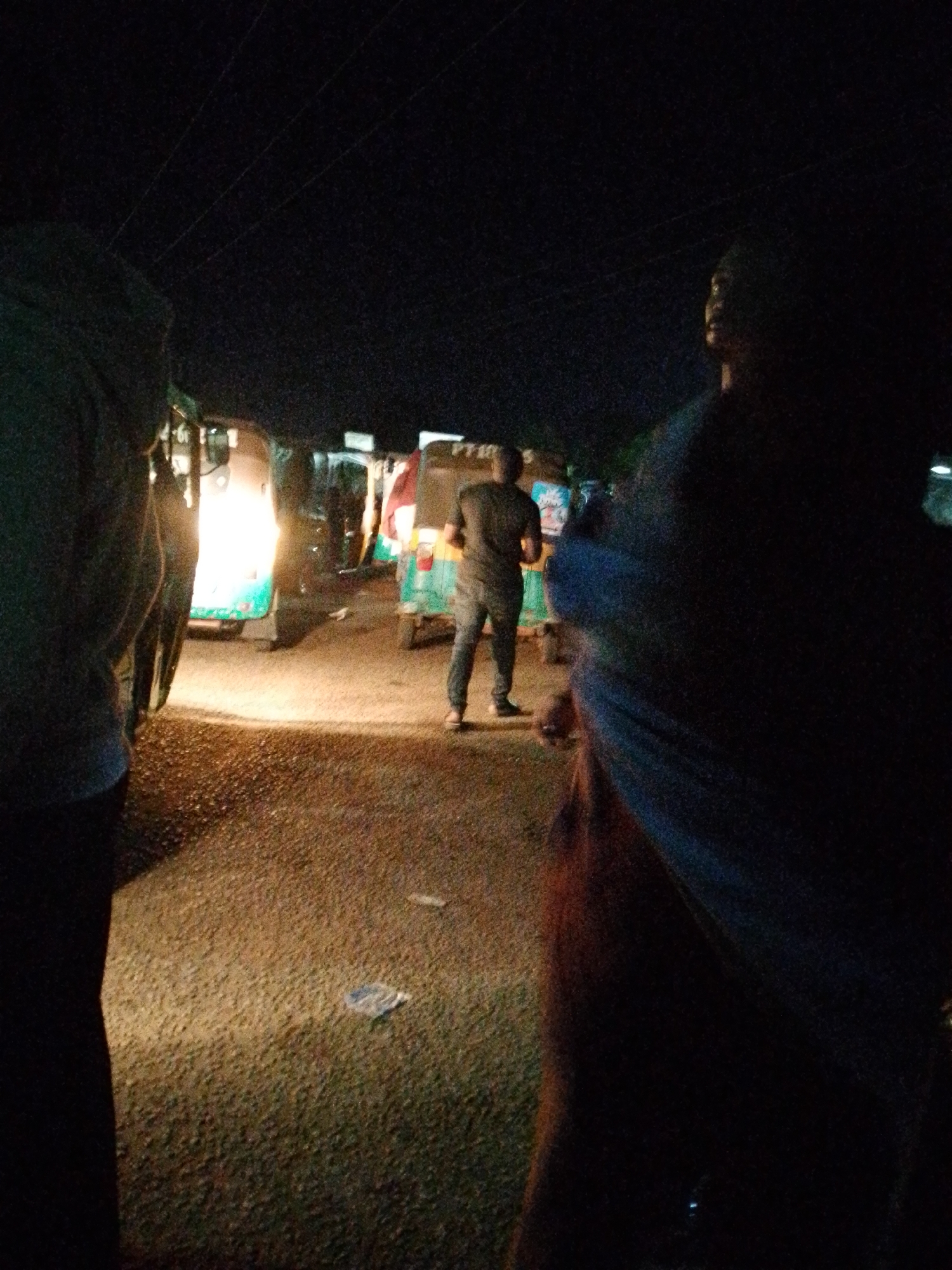 The image shows a nighttime scene in Bukuru, Nigeria. Several auto-rickshaws, also known as tuk-tuks, are parked on a road, with at least one having its headlight illuminated. A man wearing a dark shirt and pants, along with shoes, stands facing away from the camera, positioned between the auto-rickshaws. The foreground shows two people, partially cut off by the frame, one on each side of the image. The road appears to be paved, and there is some litter scattered on its surface. The overall environment is dark, with artificial lighting coming from the tuk-tuk headlights and possibly other unseen sources. Overhead are powerlines.