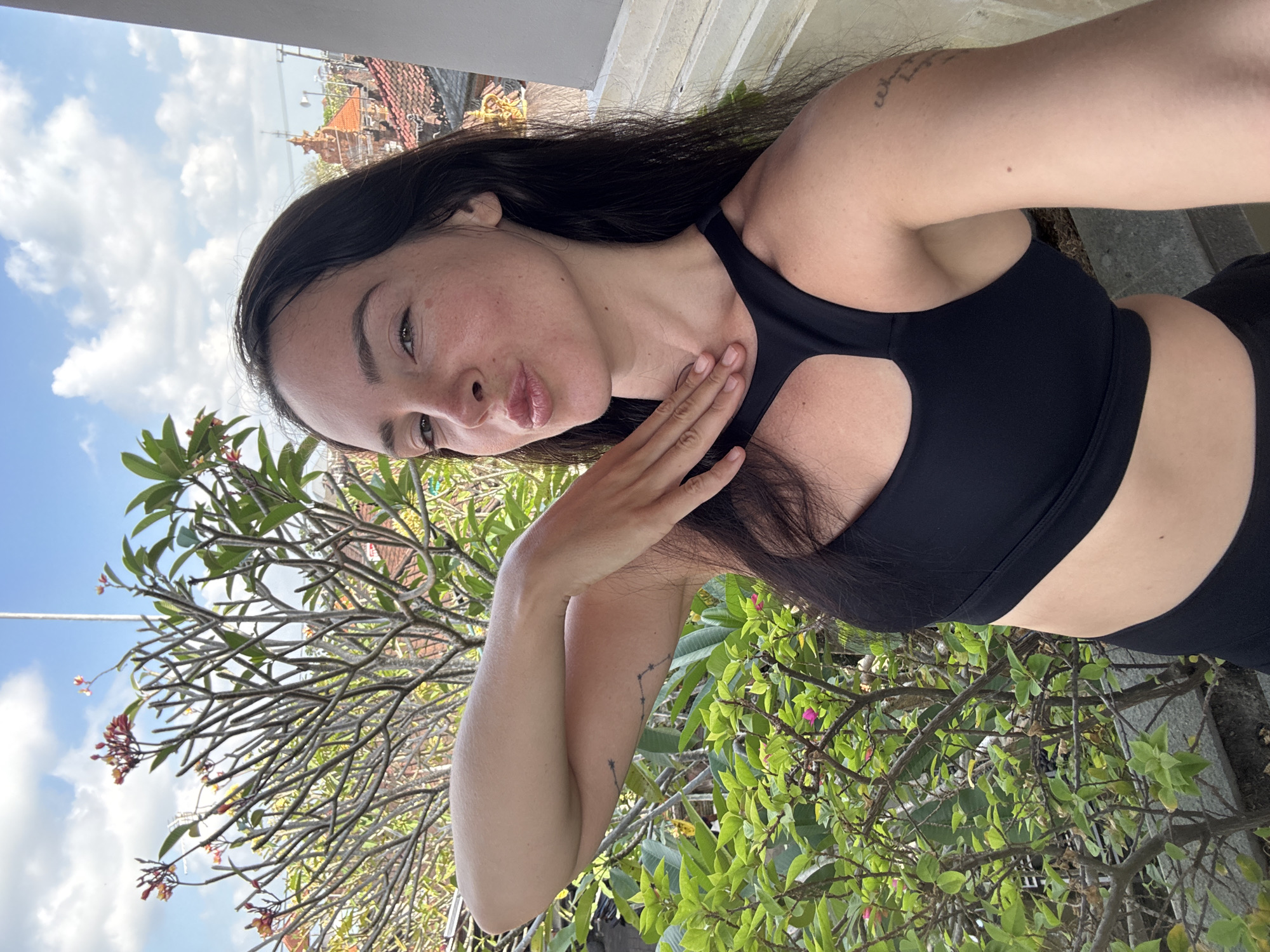 A woman in a black sports bra and black leggings is taking a selfie on a balcony in North Kuta, Indonesia. She is leaning towards the camera with her lips puckered and her left arm raised, hand resting on her neck. She has long, dark hair and fair skin with some freckles. In the background, there is lush green foliage, a tree with pink flowers, and rooftops of buildings with traditional Indonesian architecture. The sky is blue with some white clouds.