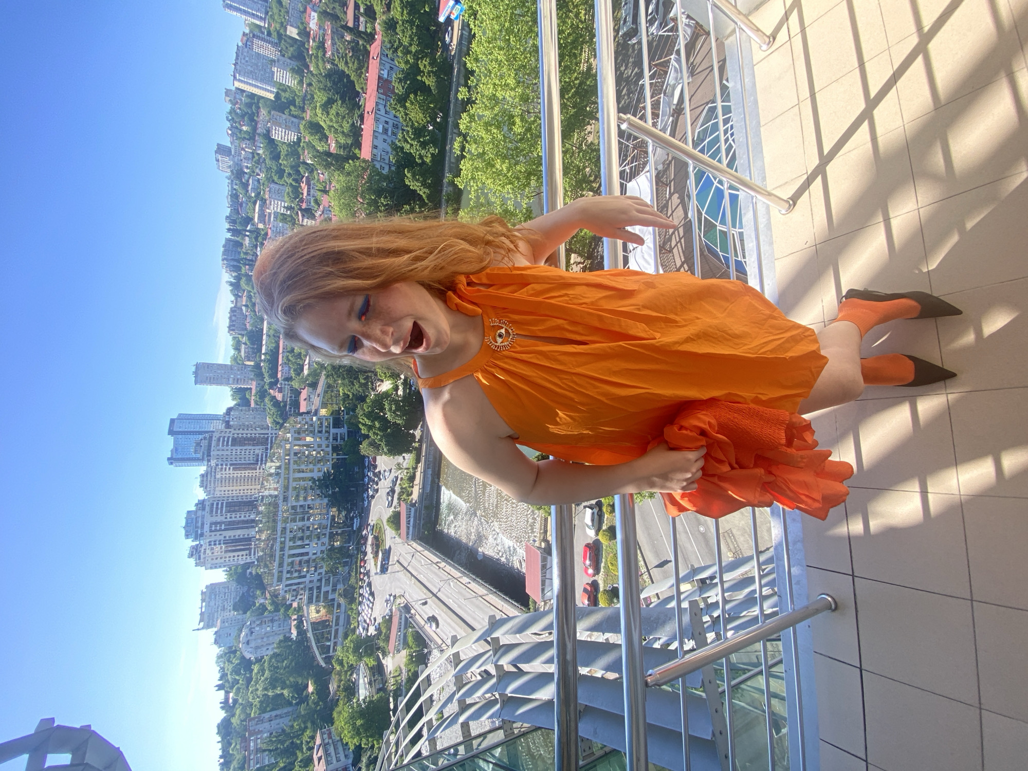 A woman with red hair and blue eye makeup stands on a balcony overlooking a city. She is wearing an orange dress and orange socks with black pointed-toe shoes. In her right hand, she holds a crumpled piece of orange fabric. The balcony has a metal railing and a tiled floor. In the background, there is a cityscape with numerous buildings and lush green trees. A road with cars and a body of water are also visible. The sky is clear and blue. The scene is likely located in Sochi, Russia, given the architectural style of the buildings and the general urban landscape.