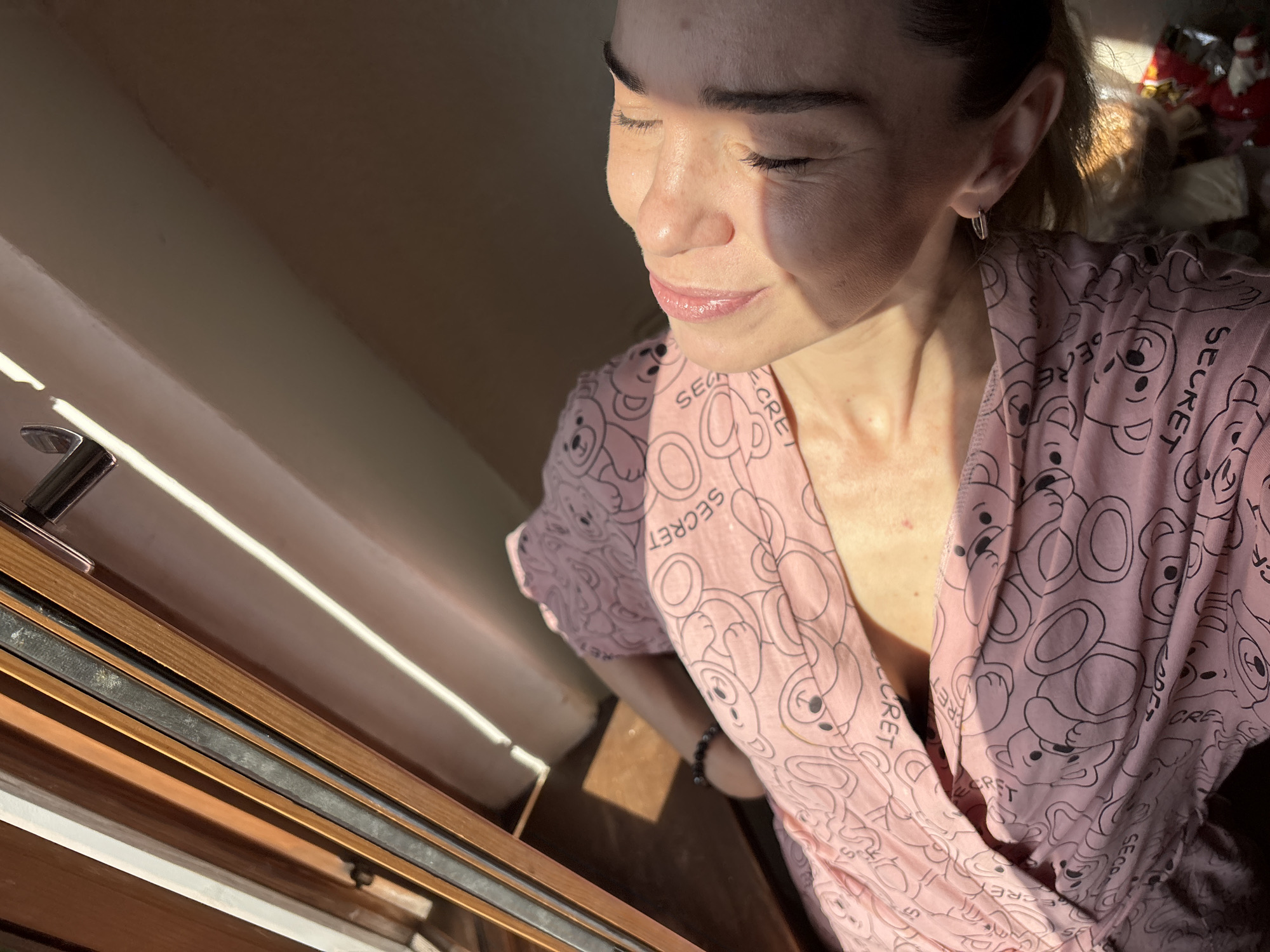 In an indoor setting, a person lies with their eyes closed, bathed in sunlight streaming from a window. The person is wearing a pink garment with a pattern of teddy bears and the word "SECRET". The sunlight highlights the person's face, which is turned upwards. The environment appears to be a room with a wooden frame and a light-colored wall. The overall scene suggests a moment of relaxation or rest. There are no identifiable landmarks that would place the location in a specific city or country.