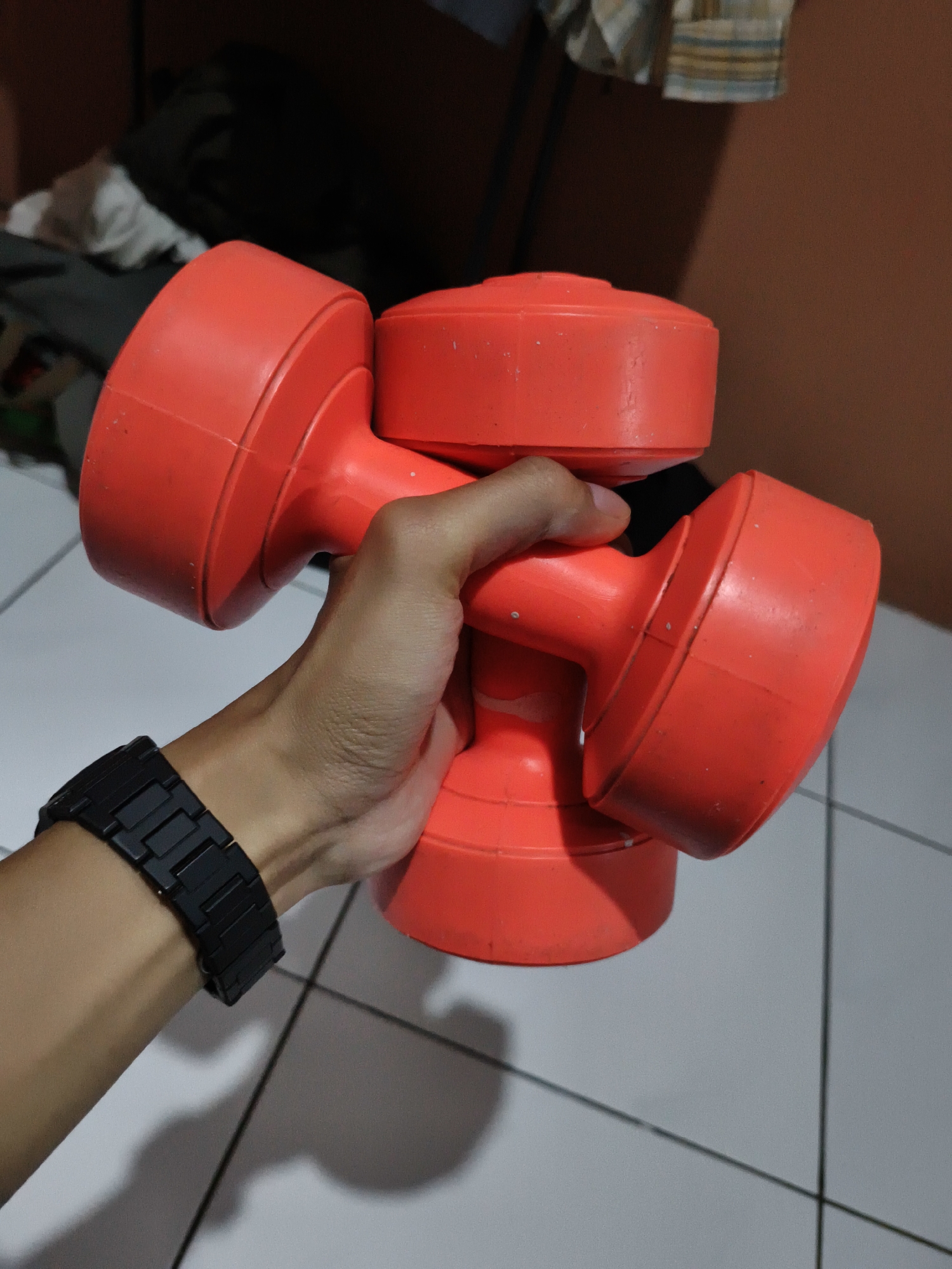 A person holds two red plastic dumbbells stacked atop one another. The person wears a black watch. The image is taken indoors on a tile floor, potentially in a home gym setting. Part of the wall is peach colored. Some items are placed in the background. It is impossible to determine if this image was taken in Pacet, Indonesia.