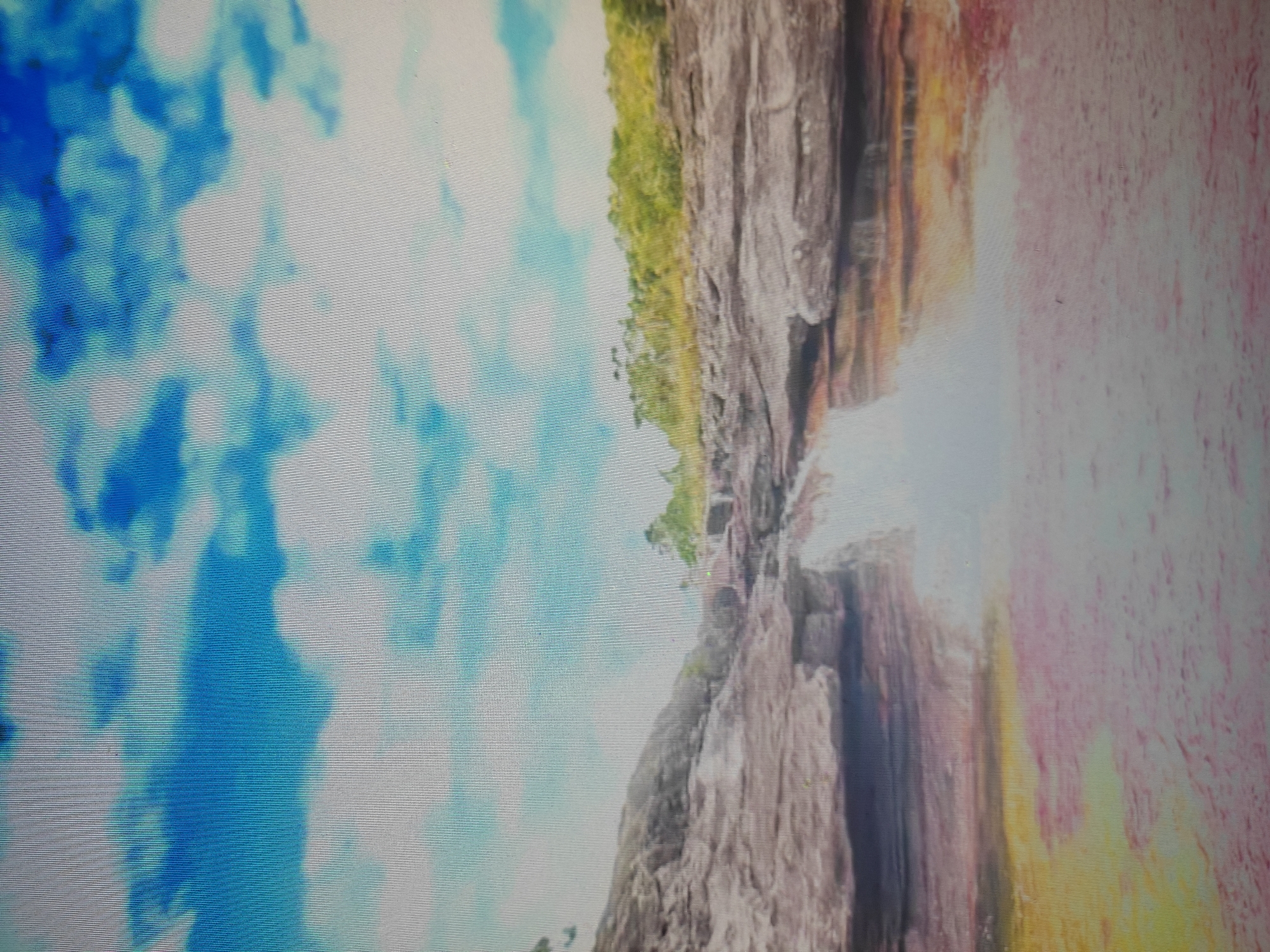 The image depicts a natural landscape with a waterfall cascading over rocky terrain. The sky above is a vibrant blue with white clouds. The water at the base of the waterfall has a pinkish-yellow hue, possibly due to mineral content or algae. Lush green vegetation covers the top of the rocks on the right side of the image, suggesting a remote or undeveloped area. The overall scene is serene and untouched.