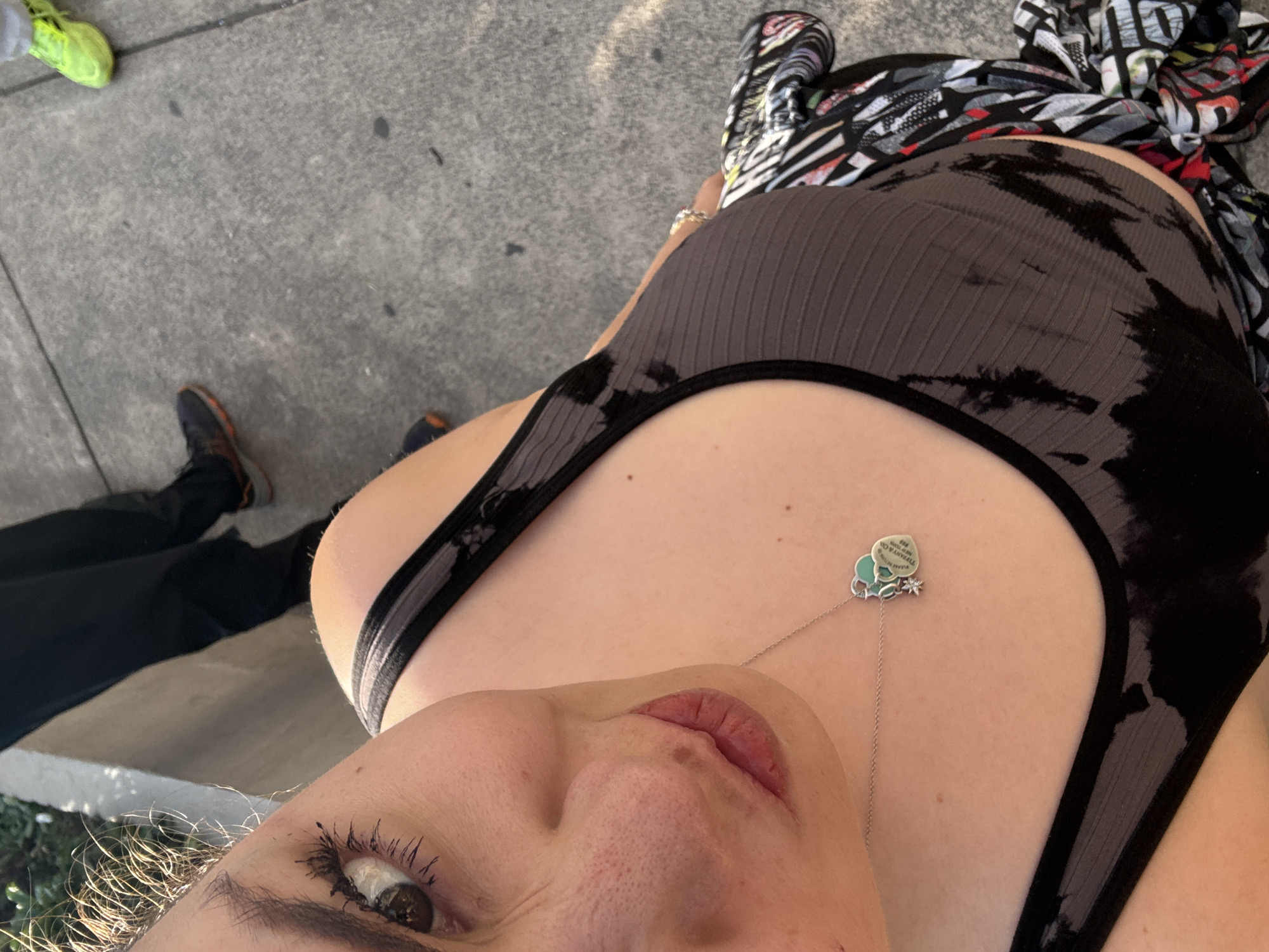 A low-angle, eye-level photograph depicts a woman wearing a gray and black tank top with a silver necklace featuring a charm. A portion of her face, including an eye, nose, and lips, is visible. Other individuals' feet, wearing shoes of varying colors, are present on the gray pavement surrounding the woman. The environment appears to be an urban setting, with a glimpse of greenery at the bottom left. A multi-colored scarf or garment is also visible adjacent to the woman. There is nothing that conclusively proves the location in the city of Mexico City, Mexico.