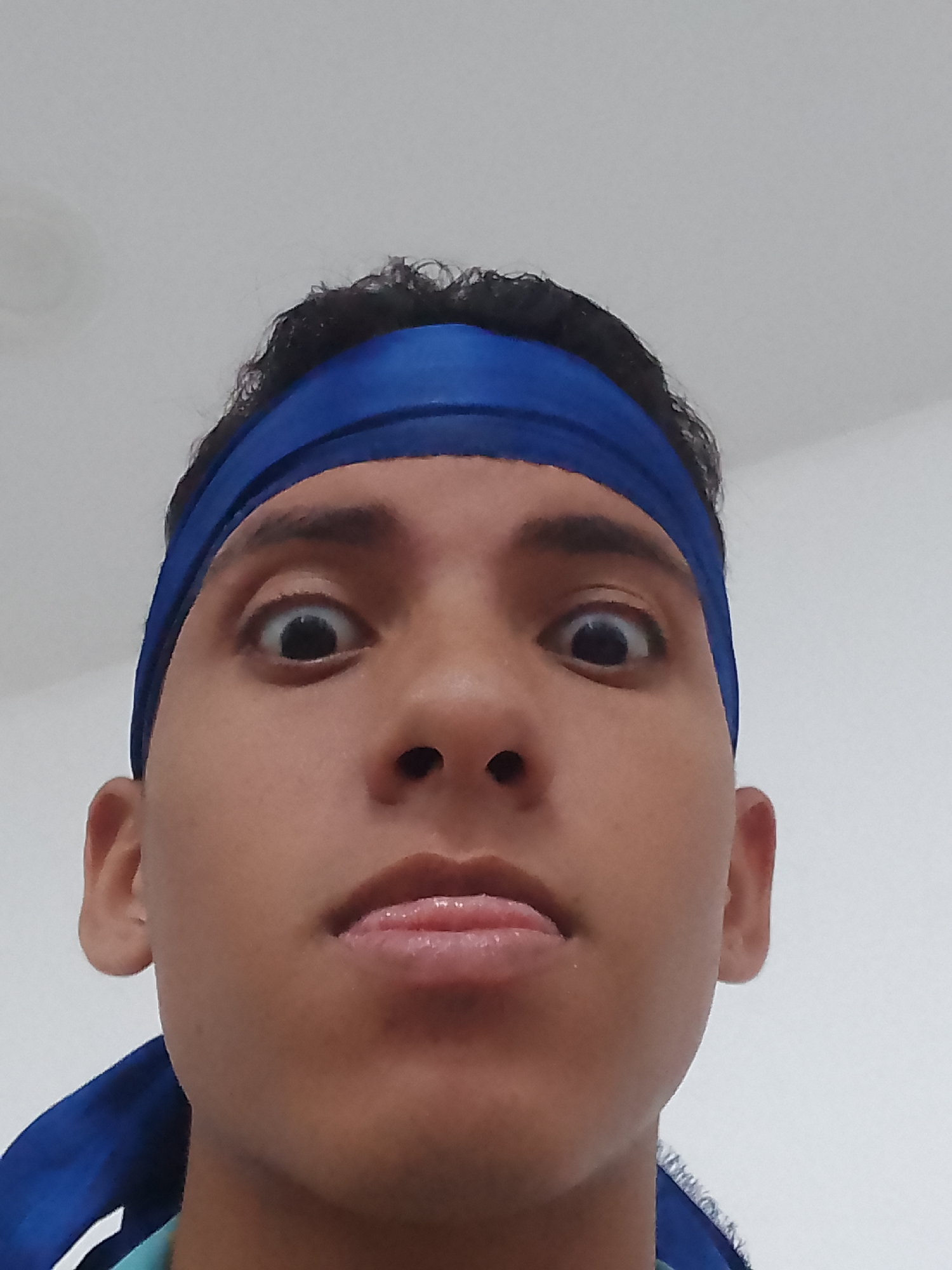 This close-up portrait, captured in Porlamar, Venezuela, features a young person, likely male, with dark, curly hair. The subject is wearing a bright blue headband and a blue and light-colored shirt or jacket, partially visible at the bottom. The person is making a humorous or exaggerated expression, with wide-open, bulging eyes and a tongue slightly protruding from the mouth. The photograph is taken from a low angle, looking up at the subject, whose face fills most of the frame. The background is a plain, light-colored surface, suggesting an indoor setting. There are no discernible location cues within the image itself beyond the provided context, nor any visible text. The lighting appears even, without harsh shadows.