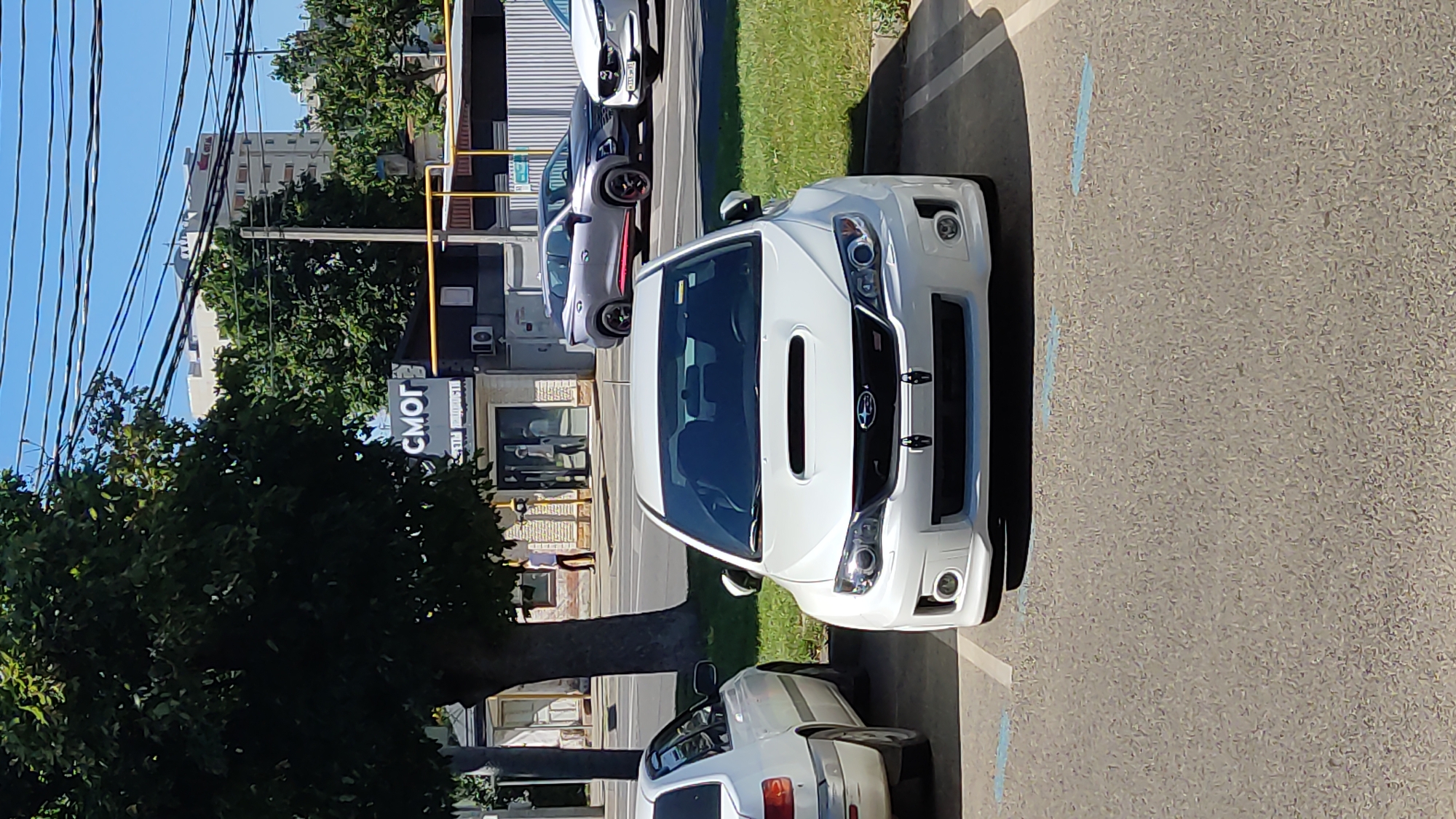 This media file is a photograph taken outdoors, likely on a sunny day given the clear blue sky and bright lighting. The scene depicts a street or parking lot with several cars. The primary focus is on a white Subaru car, seen from the front, parked in a marked parking space. Another white car is partially visible behind it. Further down the street, a light purple and a white car are also parked. In the background, buildings and trees are visible, suggesting an urban or suburban setting. A sign with Cyrillic text, reading "СМОГ", is prominently displayed on a building to the left of the frame. The image appears to have been taken around midday.