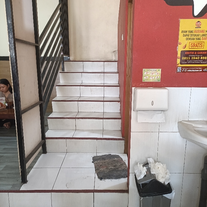 The image depicts a set of stairs in a tiled hallway within a building located at latitude -6.962 and longitude 110.256 in the city of Kaliwungu, Indonesia. The stairway has seven white tiled steps with red trim on each step, leading to an upper floor. A black metal railing runs along the side of the stairs, and a wooden partition is to the left. In the background, a woman sits at a table to the left. A white paper towel dispenser is attached to the wall to the right of the stairs, and there is a trash can with used paper towels in it. The wall to the right is tiled and painted red, with a white advertisement on it. The floors are white tiled.