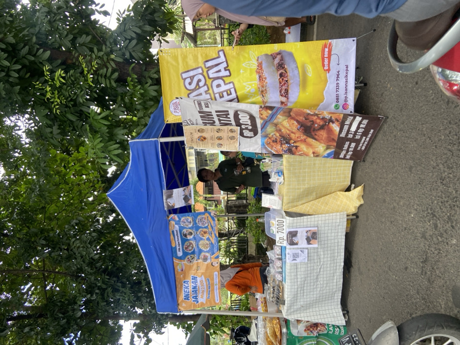 The image shows a street food stall in Kecamatan Sidoarjo, Indonesia, located at latitude: -7.442, longitude: 112.705. The stall is set up under a blue canopy tent. The vendor is a man wearing a black shirt and standing behind a table displaying various types of food, including fried items and pastries. A sign in front of the table indicates a price of Rp. 7000. A woman wearing a yellow shirt is standing to the left of the table, and a woman wearing a hijab is standing to the right of the table. The table is covered with a checkered yellow and white tablecloth. A large yellow sign advertising “Bakso Kepal” is positioned beside the table. It displays a phone number, an Instagram handle, and an order form. There is a motorcycle parked in front of the stall. Trees and other buildings can be seen in the background. The scene appears to be a typical street food market in Indonesia.