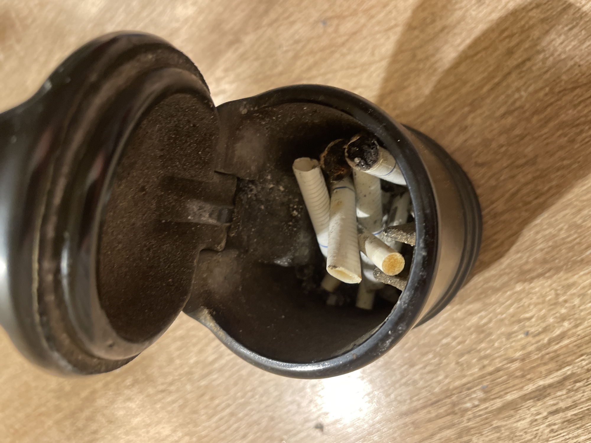 The image shows an open, black ashtray filled with several extinguished cigarette butts. The ashtray is sitting on a light-brown, wood-grain surface. There are no people visible in the image. The location in the city of Moscow, Russia, is unknown.