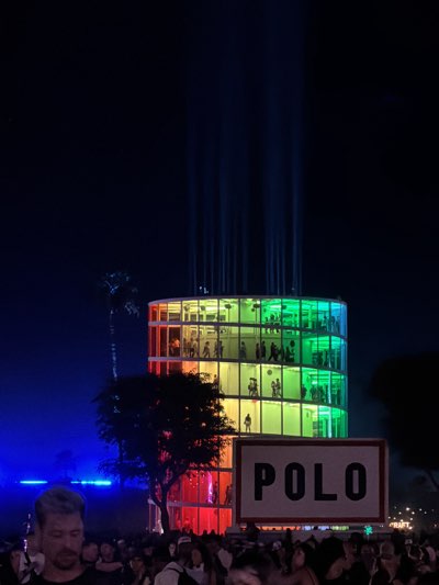 The image shows a cylindrical, multi-story building illuminated with a rainbow gradient of colors (red, orange, yellow, green) at night. The building has multiple people visible inside on each floor. Above the building, there are numerous vertical beams of light projecting upward. In the foreground, a sign with the word "POLO" is visible. A crowd of people is gathered in front of the sign and the building. The scene appears to be taking place outdoors at night in Indio, California, United States.