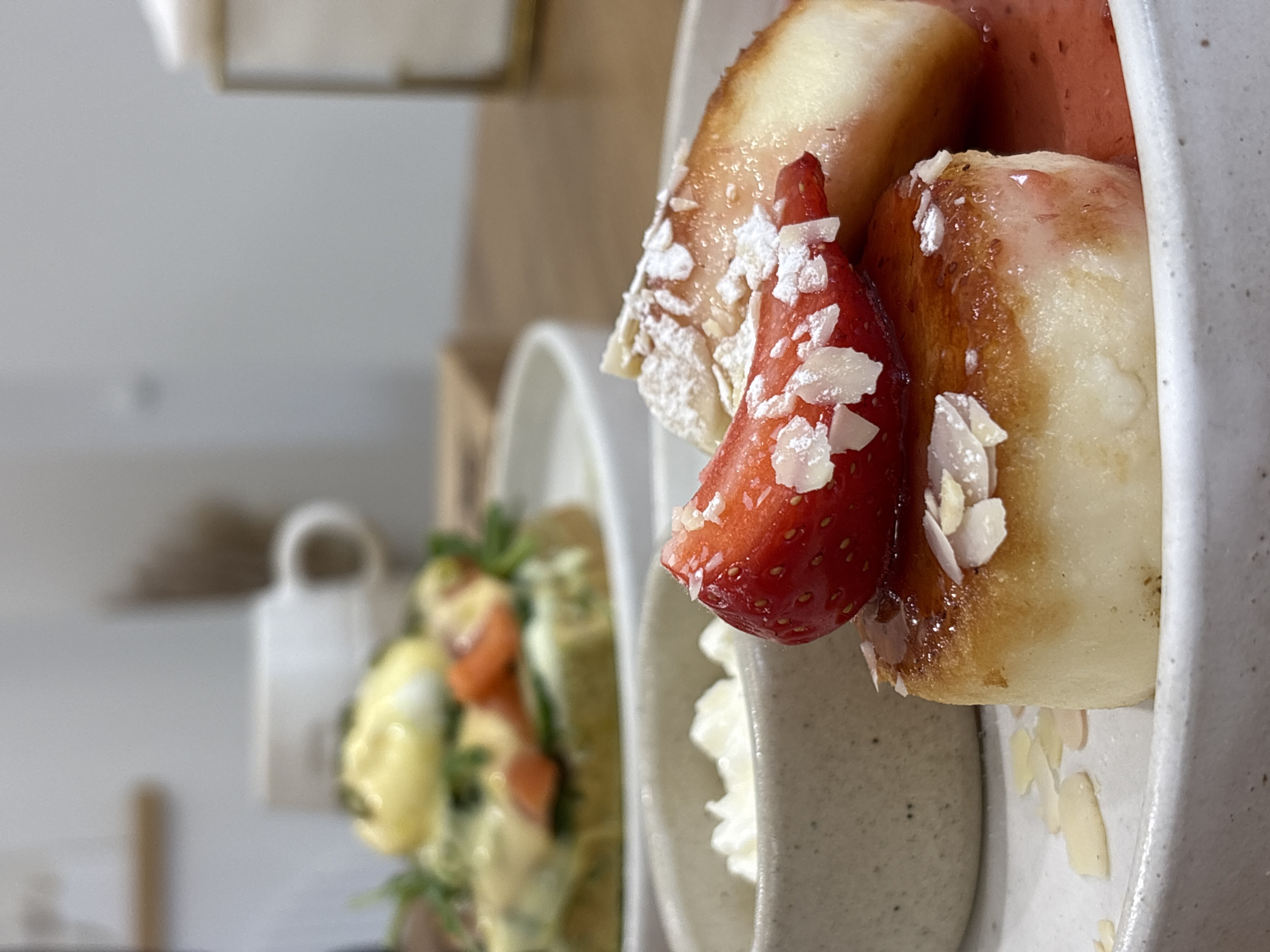 A close-up shot features a dessert on a plate. The dessert comprises what appears to be fried dough portions, a red strawberry, white whipped cream or cheese, and thin almond slices. The dessert components are coated with a translucent red sauce, possibly a berry-based syrup. The plate is made of speckled ceramic with a textured, uneven rim.
In the blurred background, another plate of food is visible, consisting of green herbs, yellow sauce, and orange pieces, possibly eggs benedict. A white mug is also visible.
The provided information does not contain enough information to confirm the location of the city of Krasnoyarsk, Russia.