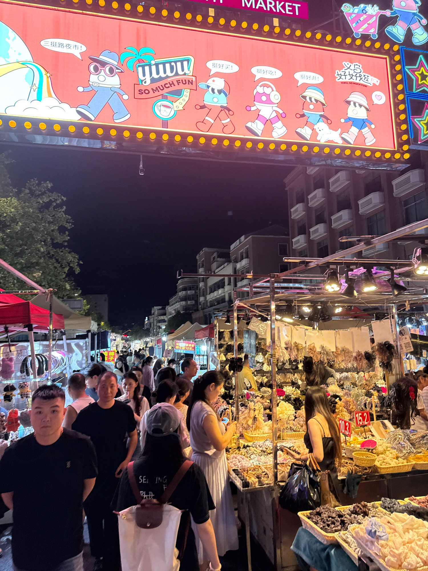 The image depicts a bustling night market in Yiwu, China, specifically located at the entrance of a market branded with "Yiwu, So Much Fun!" A large, illuminated sign above the market features cartoon characters in a playful, celebratory manner, with Chinese text that translates to "Yiwu Night Market!" Below the sign, rows of market stalls are visible, illuminated by various lights and filled with diverse merchandise, including hair accessories, clothing, and other small consumer goods. Numerous people are seen browsing the stalls and walking through the crowded pathways, indicative of a lively evening shopping experience. The overall scene is characteristic of a vibrant street market in Yiwu, a city known for its extensive wholesale markets and commercial activity.