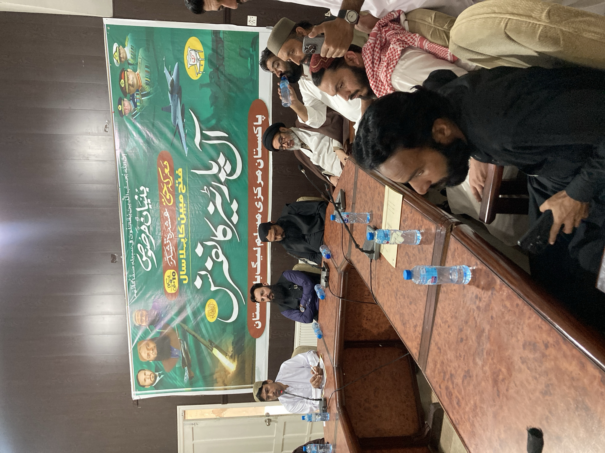 The image depicts a gathering of men in Quetta City Tehsil, Pakistan, seated around a large wooden conference table. Several individuals are visible, some in traditional Muslim attire and others in casual clothing. Water bottles are scattered across the table, and microphones suggest a formal meeting or press conference. A large banner is displayed in the background featuring images of military figures, a fighter jet, and Arabic text. The banner relates to "Pakistan Central Muslim League".