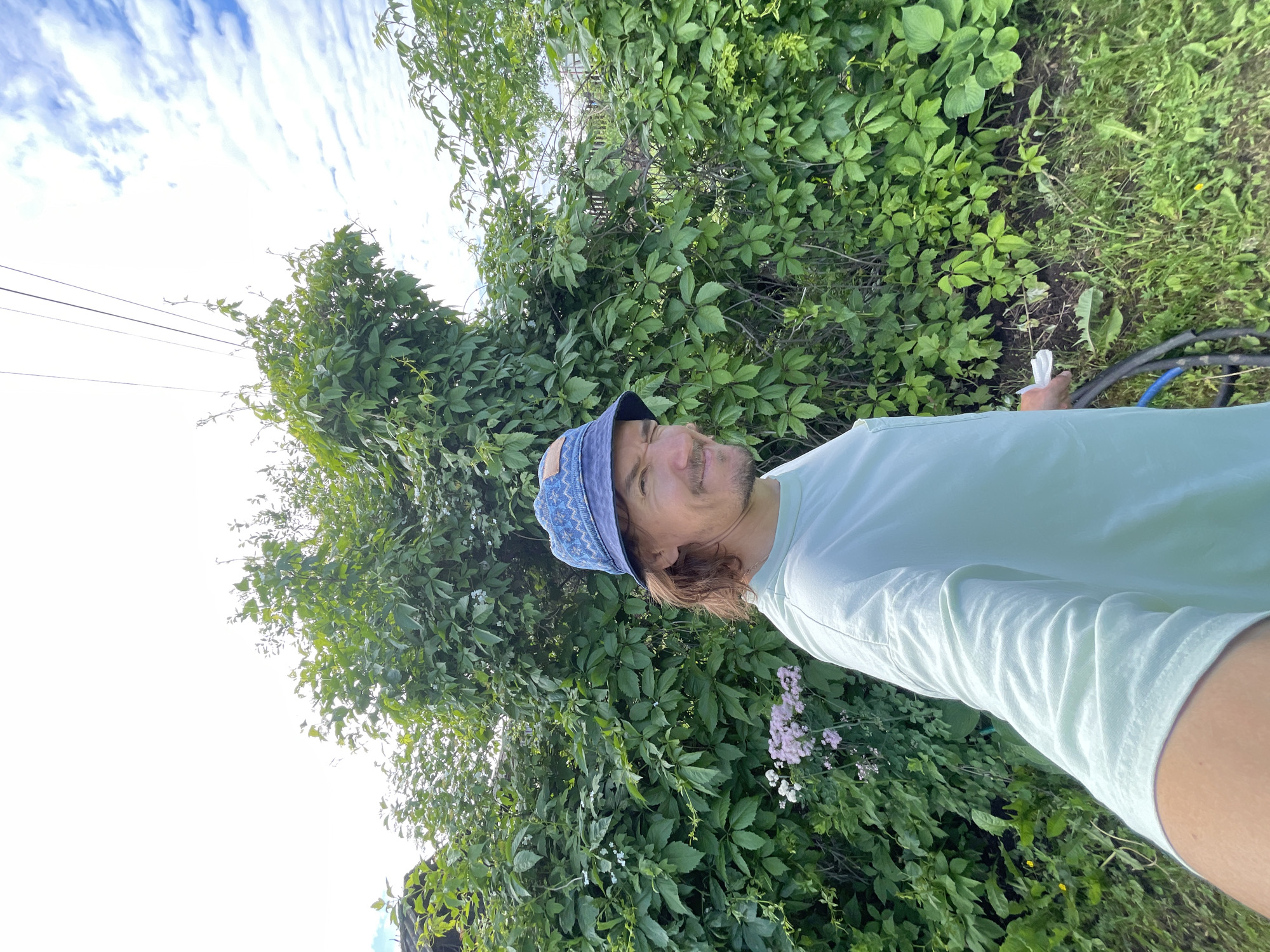 A male individual is depicted in a selfie taken outdoors in Unknown, Unknown. He is positioned in the mid-ground, slightly right of center, looking towards the camera with a wink and a slight smile. He has medium-length brown hair, a light mustache, and stubble. He is wearing a blue bucket hat with a patterned design and a light mint green long-sleeved t-shirt. His right arm, holding the camera, is visible in the lower left corner of the frame. His left hand is seen near his hip, holding a small white object.
The background consists primarily of dense green foliage, appearing as a thick wall of leaves behind and to the sides of the subject. On the far left, partially obscured by foliage, a dark, possibly corrugated roof is visible. A cluster of light pinkish-purple flowers is present in the lower left, between the subject and the foliage. In the upper central and right portions of the background, the sky is visible, mostly bright white with patches of light blue and scattered stratocumulus clouds on the right. Three dark, thin power lines extend horizontally across the upper part of the image, emerging from the foliage on the left and crossing into the sky on the right. The ground in the foreground consists of green grass with some weeds and patches of bare earth. A coiled black hose with a visible blue section lies on the ground to the right of the subject's lower body.