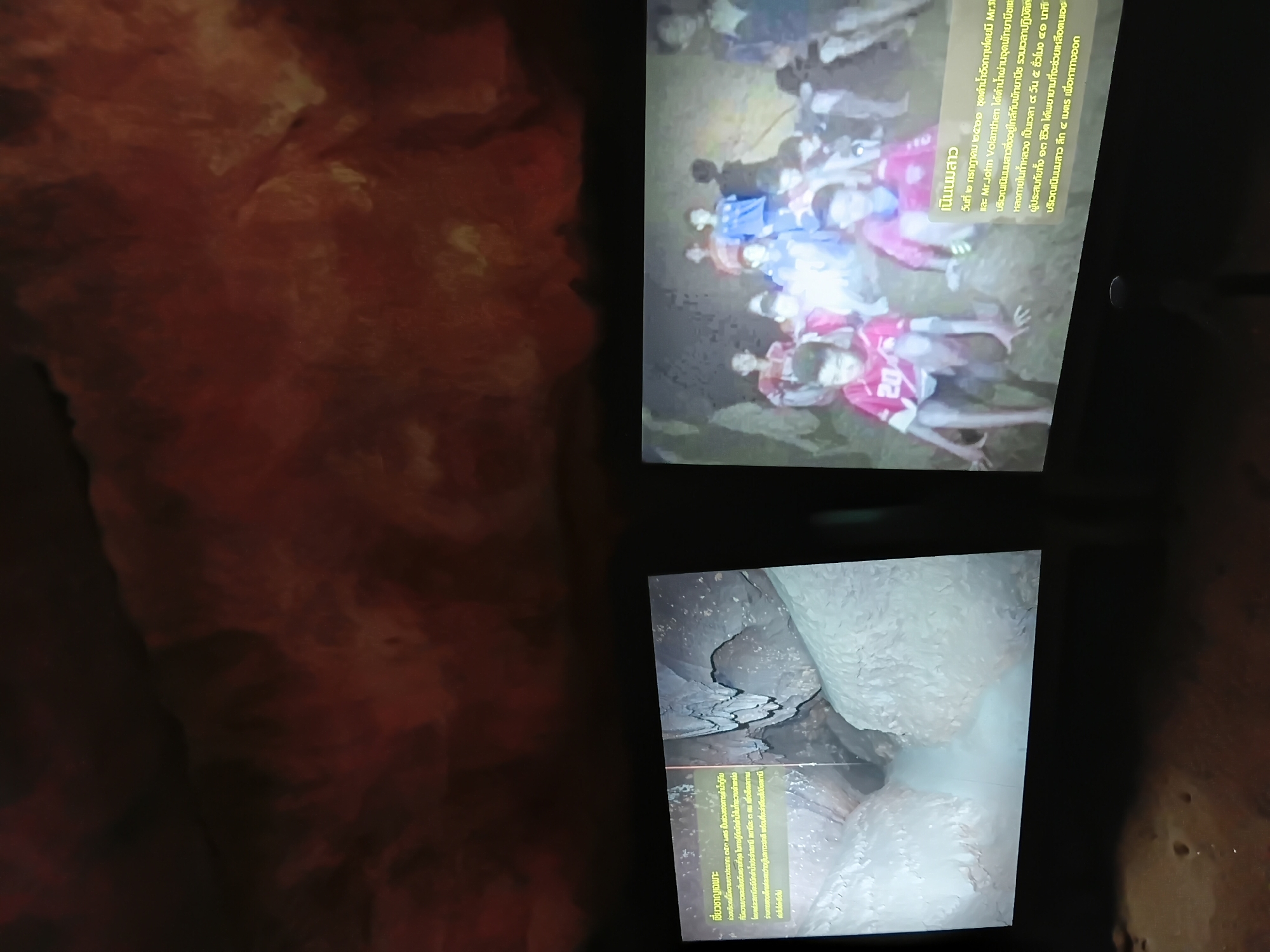 This is a photograph taken at the Tham Luang cave complex in Mae Sai District, Thailand. The image displays two screens with projected content. The left screen shows a close-up view of the cave's interior, featuring water flowing through a narrow passage between large rock formations. The right screen displays a group of young boys, presumably survivors of the cave rescue, in what appears to be the cave environment. Text in Thai is visible on both screens, providing context about the cave and the rescue event. The background of the photograph shows a dimly lit, textured wall, possibly part of the cave's natural environment or a display setting.