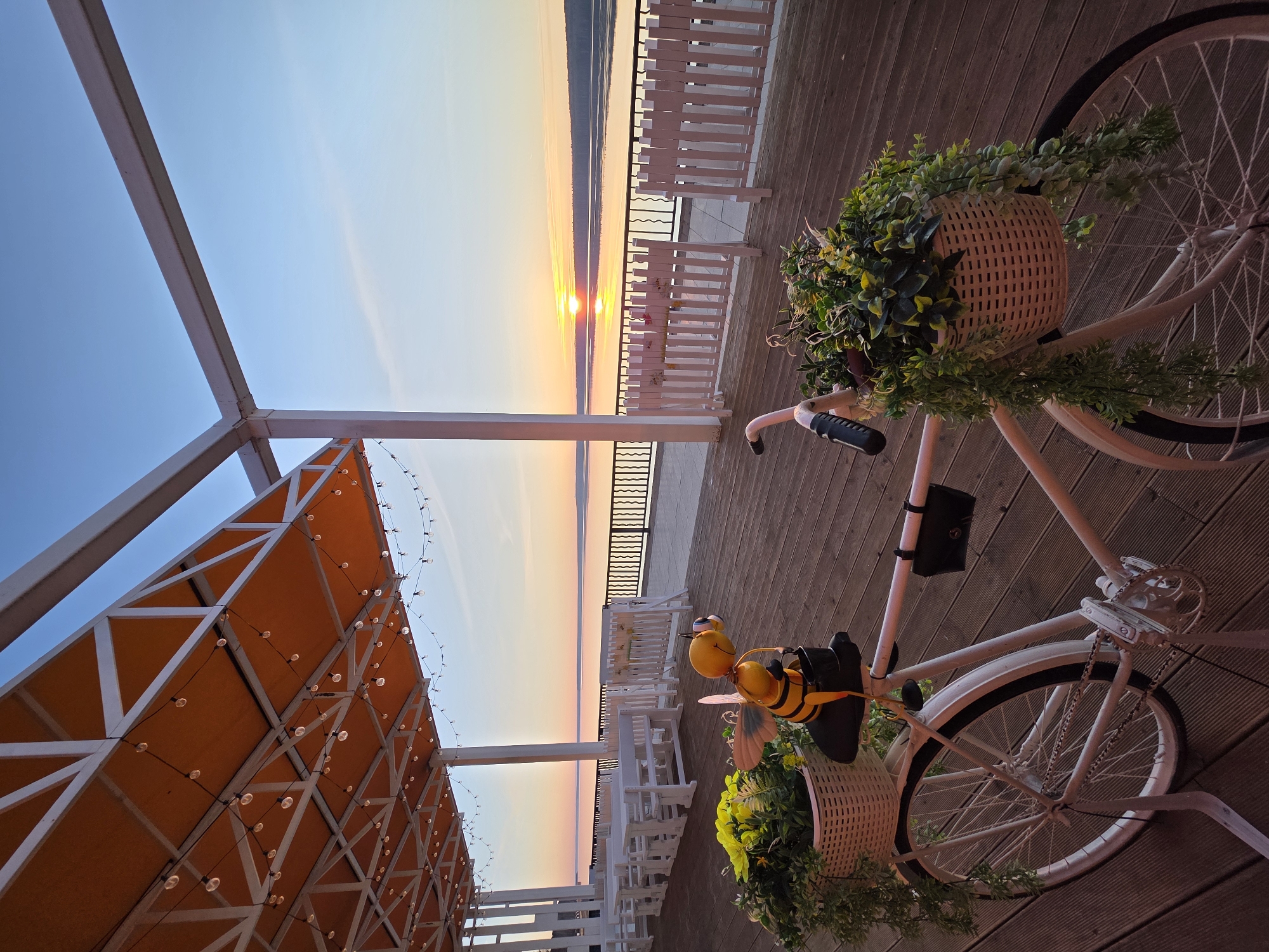 Here's a detailed description of the image:
**Location:** The image appears to be taken in the city of Chaikovsky, Russia, based on the presence of a bicycle and decorations similar to what is found in the city.
**Subjects and Objects:**
* **Bicycle:** A white bicycle is prominently featured in the foreground. It has baskets attached to the front and back, filled with plants.
* **Decorations:** There is a decorative bumblebee attached to the front of the bicycle.
* **Setting:** The bicycle is placed on a wooden deck or patio. A white railing lines the edge of the deck, with glimpses of a body of water (possibly a river or lake) and the sunset visible in the background.
* **Pergola:** A partial view of a pergola-like structure is seen on the left side of the image. It has an orange awning or covering and is adorned with string lights.
* **Furniture:** There is a line of white tables and chairs in the background.
**Environment:**
* **Lighting:** The image is taken during sunset, as evident from the warm colors in the sky.
* **Atmosphere:** The scene evokes a tranquil, possibly romantic ambiance with the sunset and decorative elements.
* **General Impression:** The setting suggests a cafe, restaurant, or other outdoor leisure area.