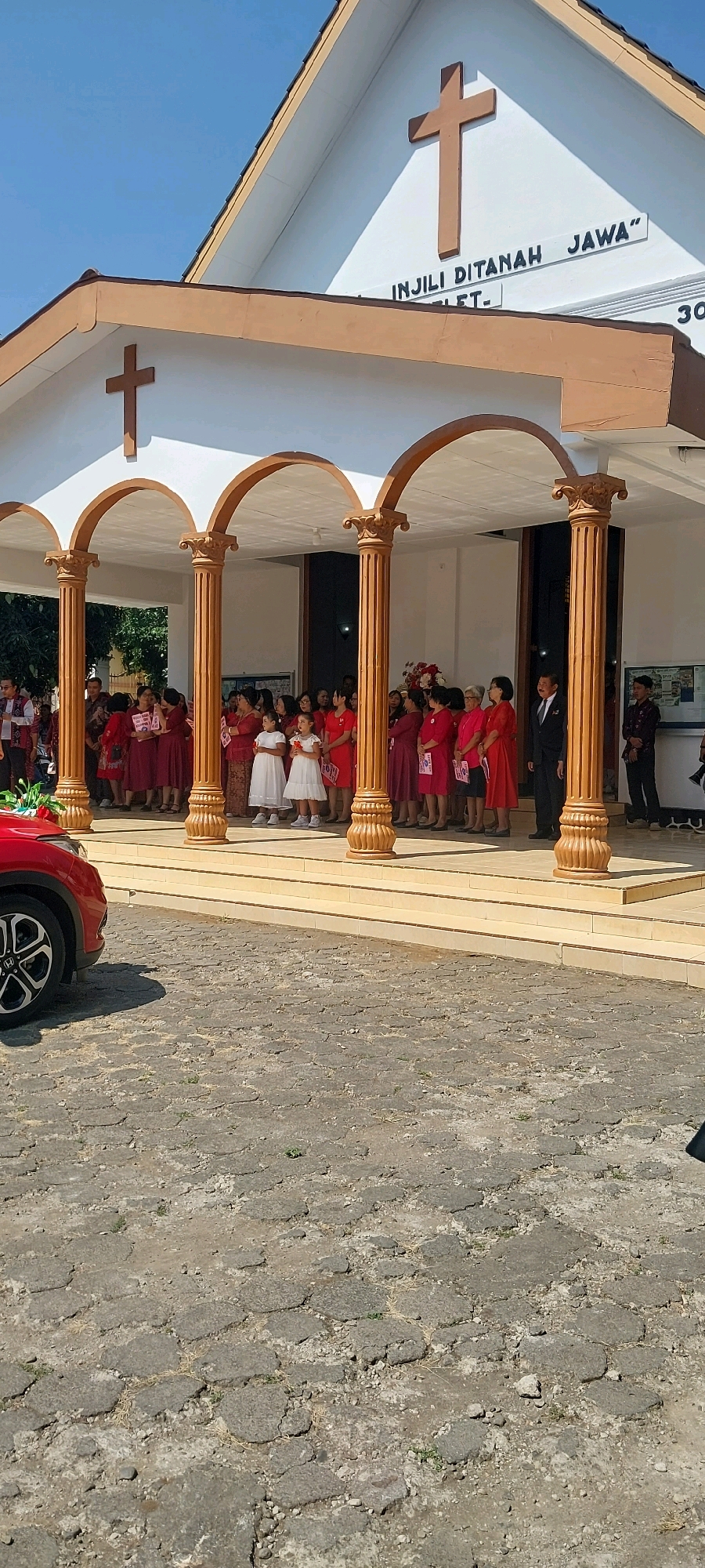 The image shows the entrance of a church in Keling, Indonesia, with a group of people gathered outside. The church has a white facade with brown trim and features a large cross at the apex of the roof. The entrance is adorned with three arched openings supported by ornate columns.
A group of women and girls dressed in red and white are standing under the arches, facing the camera. Two young girls in white dresses are at the forefront of the group, holding what appear to be prayer books or pamphlets. The women are also holding similar items. Some of the women are wearing red dresses, while others are in darker red or maroon attire.
To the right of the group, a man in a dark suit is standing, looking towards the right. Further to the right, a man in a dark shirt is partially visible.
In the foreground, a red car is parked on a cobblestone driveway. The car's left front wheel and fender are visible. The cobblestone pavement is uneven and shows signs of wear and tear, with some grass growing through the cracks.
The sky above is clear and blue, indicating a sunny day. The overall scene suggests a religious gathering or event taking place at the church.