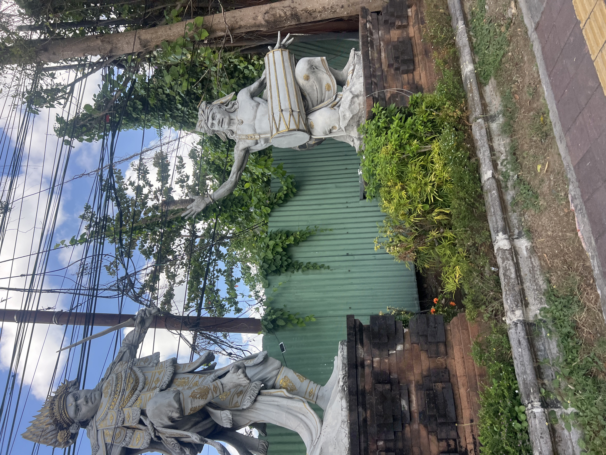 The image depicts two statues in Denpasar, Indonesia. On the left, a large stone statue of a warrior with a crown and elaborate armor stands holding a sword. On the right, another stone statue of a man with a headdress is shown playing a drum. Both statues are positioned on raised platforms, with a green corrugated metal fence behind them. Lush green vegetation surrounds the statues, and overhead, a tangle of electrical wires crisscrosses the sky, which is partly cloudy. The foreground shows a paved walkway and overgrown grass. The style of the statues suggests a Balinese artistic influence, commonly found in public spaces and temples throughout Denpasar.