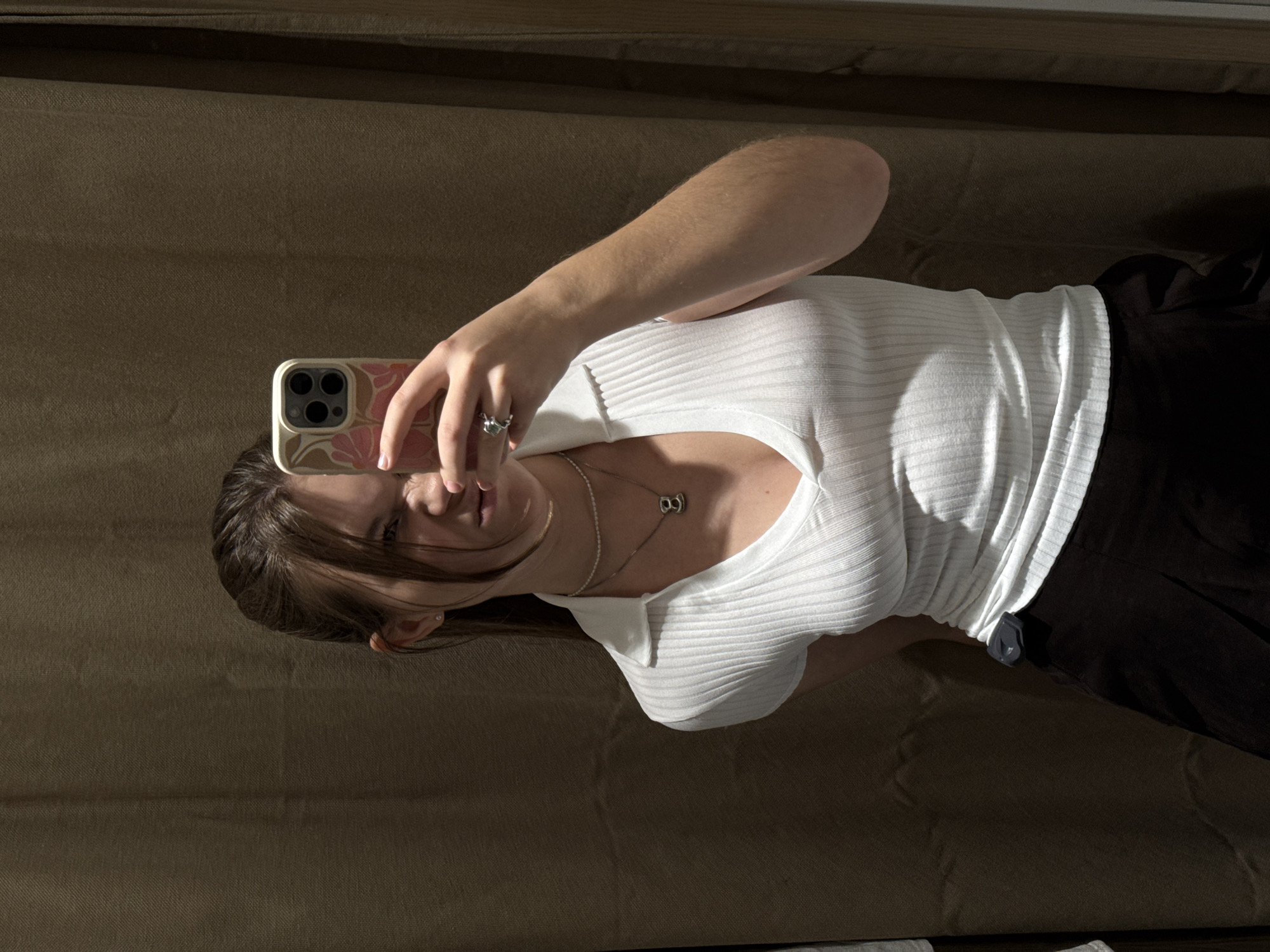 A person is taking a selfie using a smartphone. The individual is wearing a white, collared, ribbed shirt and black pants. They have light skin, brown hair, and are wearing a necklace with a pendant. They are holding the phone with their right hand, where they wear a ring. The phone has a patterned case. The background consists of a textured, possibly upholstered, surface, suggesting a couch or wall covering. There is no visible information to indicate the location is Belgrade, Serbia.