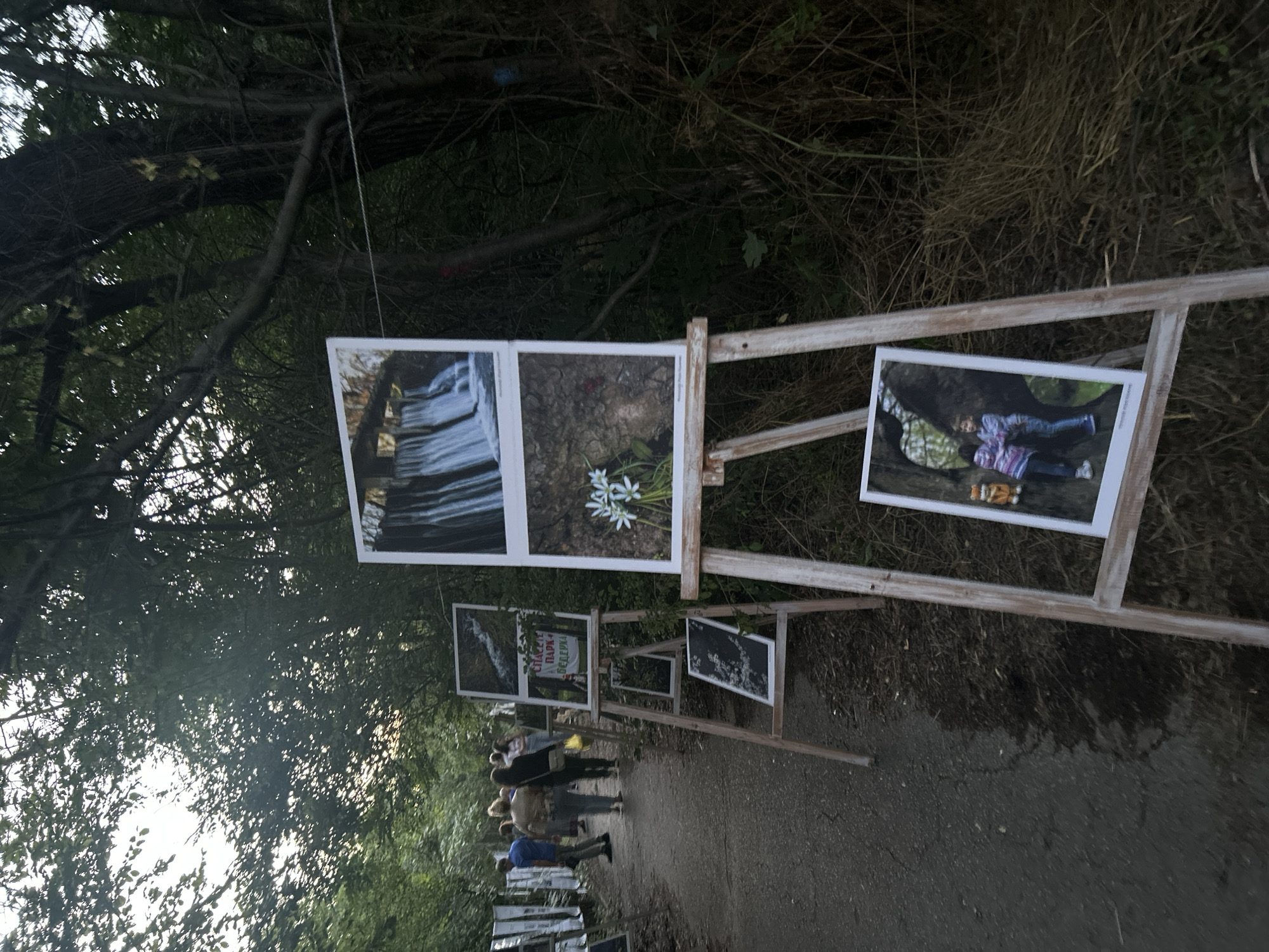 The image shows an outdoor art exhibition located on a paved path with trees and vegetation on either side. Several photographs are displayed on wooden easels. The photographs depict natural scenes, including a waterfall, a flower, and two children posing. There are a few people walking along the path, appearing to be visitors to the exhibition. The lighting suggests it is late afternoon or early evening.