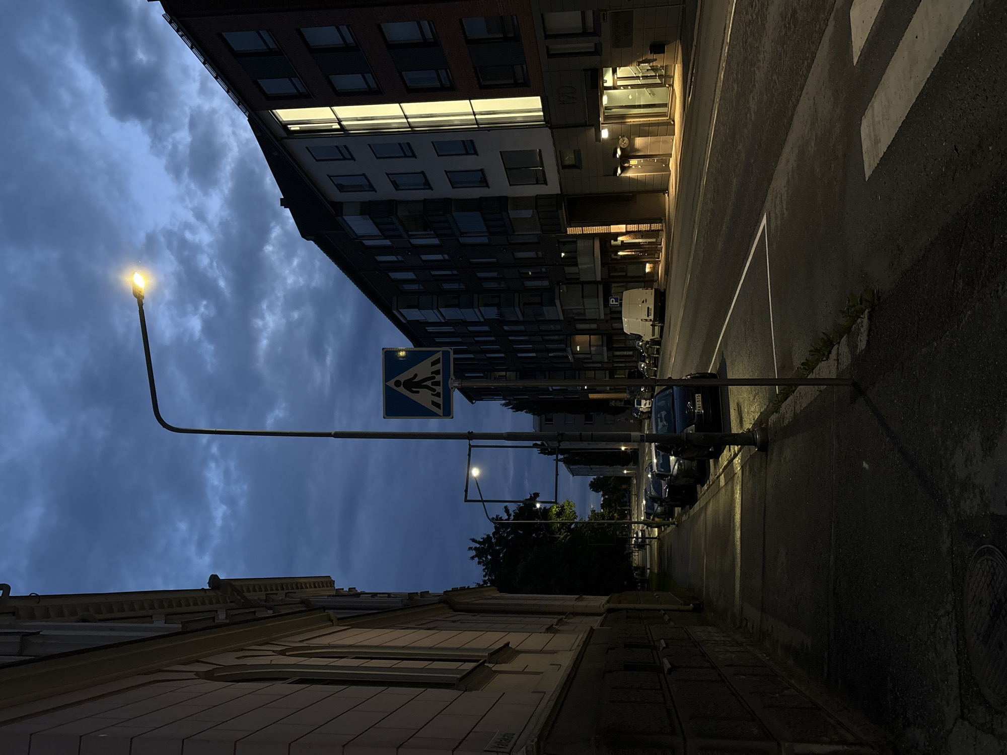 The image depicts a street scene in Pori, Finland, at dusk or early night. The sky is a deep blue with dark clouds. A streetlamp illuminates a pedestrian crossing on the right side of the image, where there are also modern apartment buildings with illuminated windows. On the left side, a building with arched windows and ornate architectural details lines the sidewalk. Cars are parked along the street, and a white van is visible in front of one of the apartment buildings. The street is paved, and a sidewalk runs alongside the buildings. The overall atmosphere is quiet and dimly lit, characteristic of an urban environment transitioning from day to night.