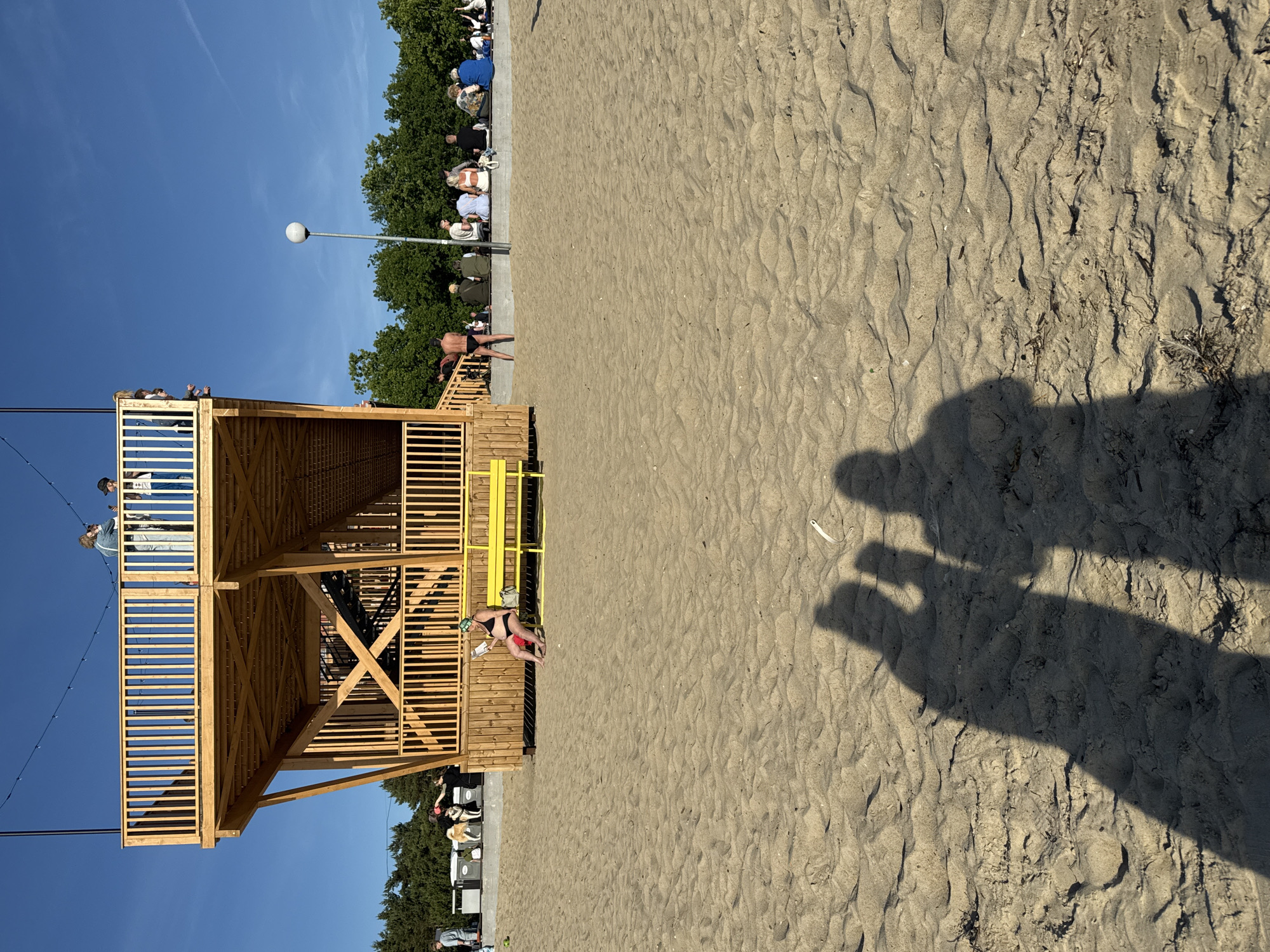 A bright, sandy beach in Tallinn, Estonia, is the setting for this daytime scene. Dominating the mid-ground is a multi-level wooden structure, possibly a viewing platform or a pavilion, with a central staircase. On the upper level, three people are visible, two of whom appear to be looking at their phones. Below, on the ground level, a woman in a black bikini sits on a yellow bench, reading. Further back, a group of people are seated on a low wall, observing the scene. A man in black swim briefs stands near the wooden structure. The beach itself is largely empty, with only a few scattered objects and the shadows of two people in the foreground. The background features a line of green trees against a clear blue sky.