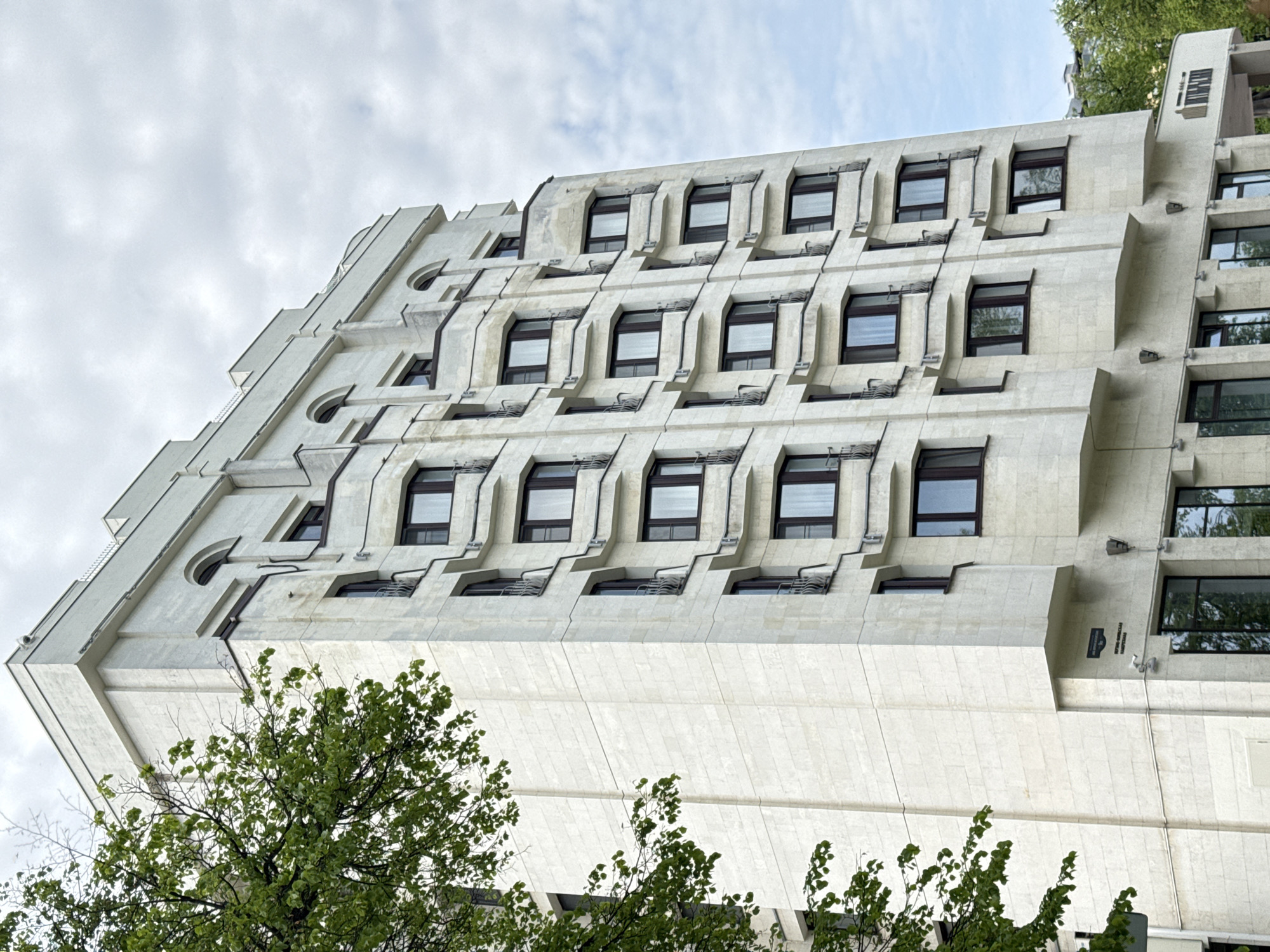 The image shows a low-angle view of the Central Bank building in Nizhny Novgorod, Russia. The building is a modernist structure with a distinctive facade featuring a repetitive pattern of recessed windows and protruding concrete or stone elements. The exterior is primarily light in color. In the foreground, leafy green trees partially obscure the lower portion of the building. The sky is overcast, with light grey clouds.