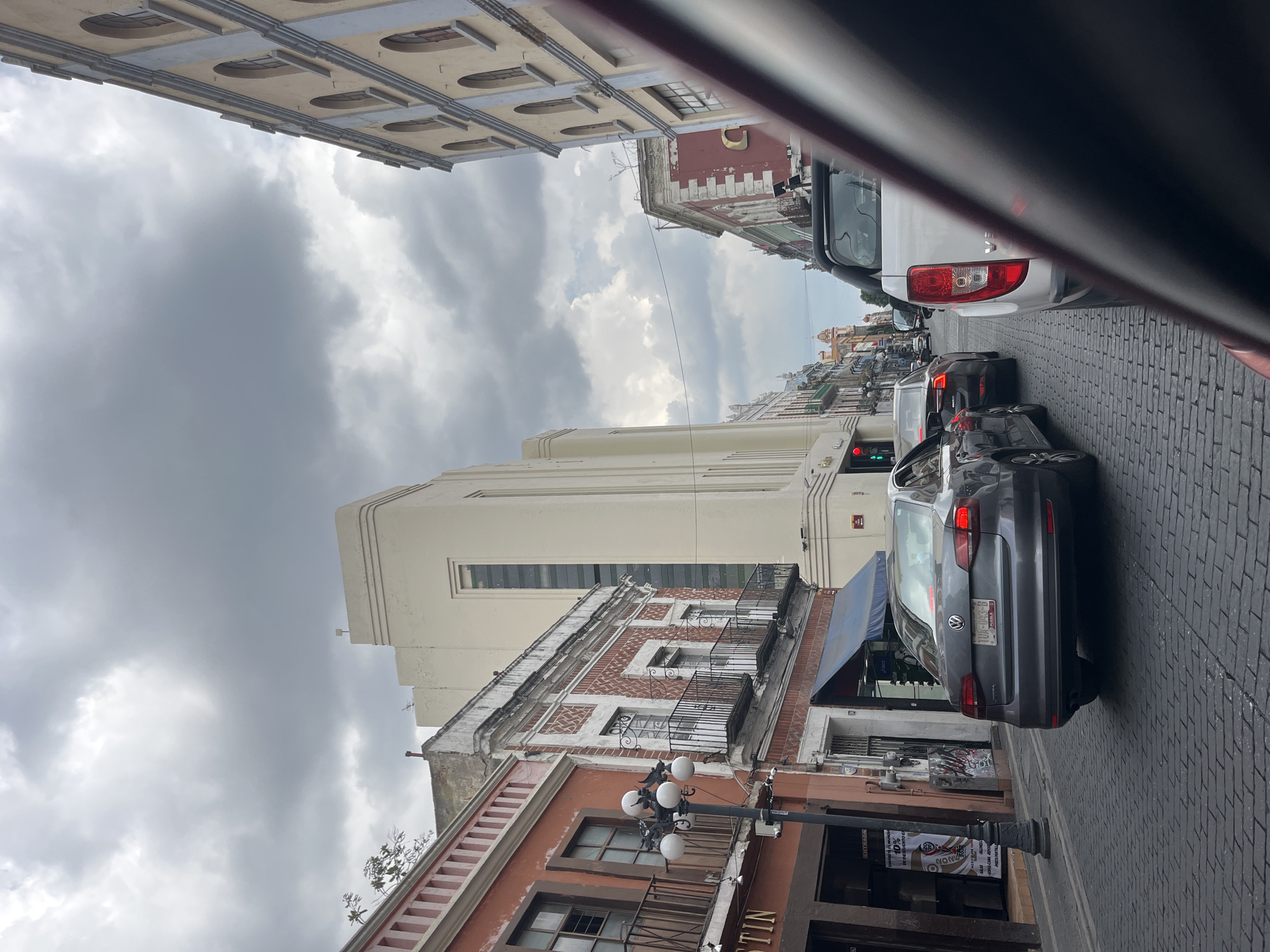 The image shows a street scene in Puebla, Mexico. The view is taken from a moving vehicle, evidenced by the blurred foreground. The street is paved with grey cobblestones, and a line of cars are stopped at a red traffic light. Buildings flank both sides of the street, showcasing a mix of architectural styles, including ornate facades with balconies. A street lamp is visible on the lower left. Overcast skies dominate the upper portion of the frame. The businesses on the street are not clear, although signage and posters are seen.