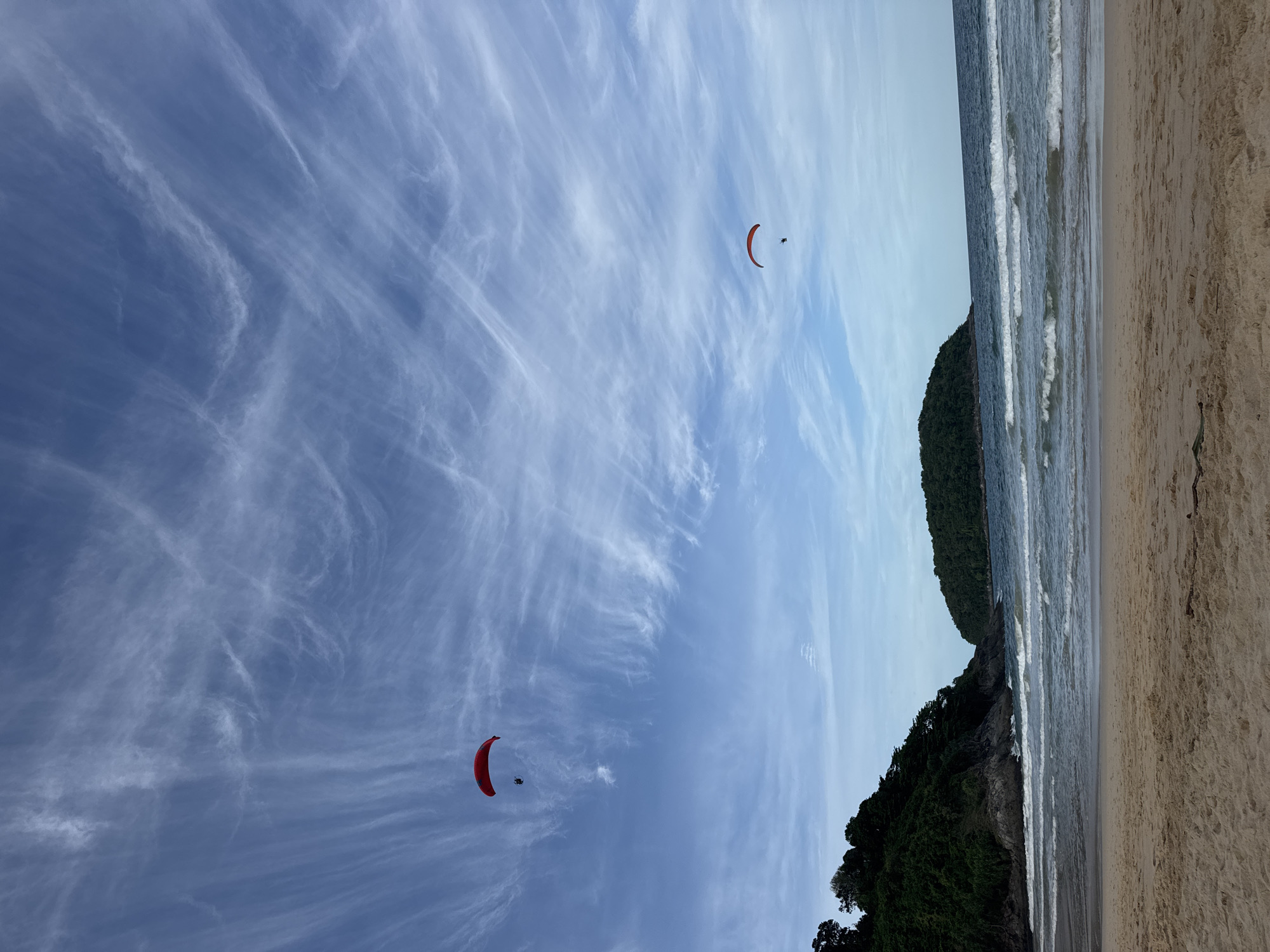 The scene captures a coastal landscape in Rawai, Thailand, featuring a wide sandy beach, ocean, verdant hills, and two paragliders under a wide blue sky.
The upper two-thirds of the image are filled with a clear blue sky, streaked with numerous thin, white cirrus clouds that create a wispy, elongated pattern across the frame. Two individuals are visible airborne under red paraglider canopies. One paraglider, larger in frame, is positioned in the upper-left quadrant, with its pilot visible below the canopy. The second paraglider, smaller and more distant, is located in the middle-right.
The mid-ground features a lush, dark green, tree-covered hill or cliff rising steeply from the water on the left side of the frame. Rocky formations are discernible at the base of this landmass where it meets the ocean. To the right of the hill, a smaller, similarly forested island is visible in the distance, separated by a stretch of water.
The lower third of the image is occupied by the ocean, which exhibits moderate waves breaking onto the sandy shore, creating white foam lines. The water displays varying shades of blue and green. The immediate foreground is a light brown sandy beach, showing subtle texture and undulations. A dark, elongated object, possibly a piece of driftwood or debris, rests on the sand in the lower-right corner.