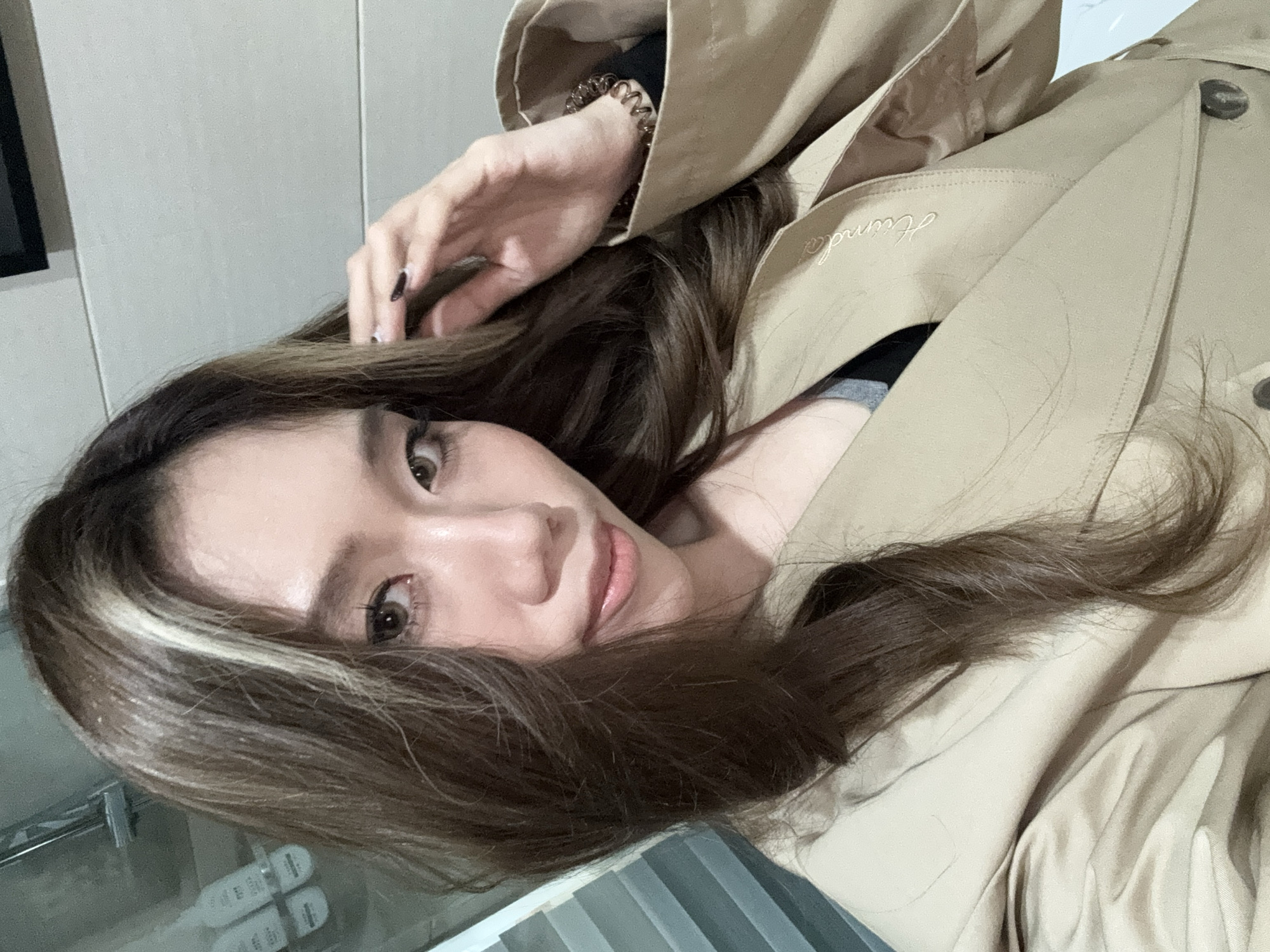 A young woman with brown hair and a blonde streak is pictured in a close-up selfie. She is wearing a tan trench coat. Part of a wrist adornment is visible on her left wrist. The background is indistinct, but appears to include a wall, a display of products, and a set of paint swatches. No information connects this to Lanzhou, China.