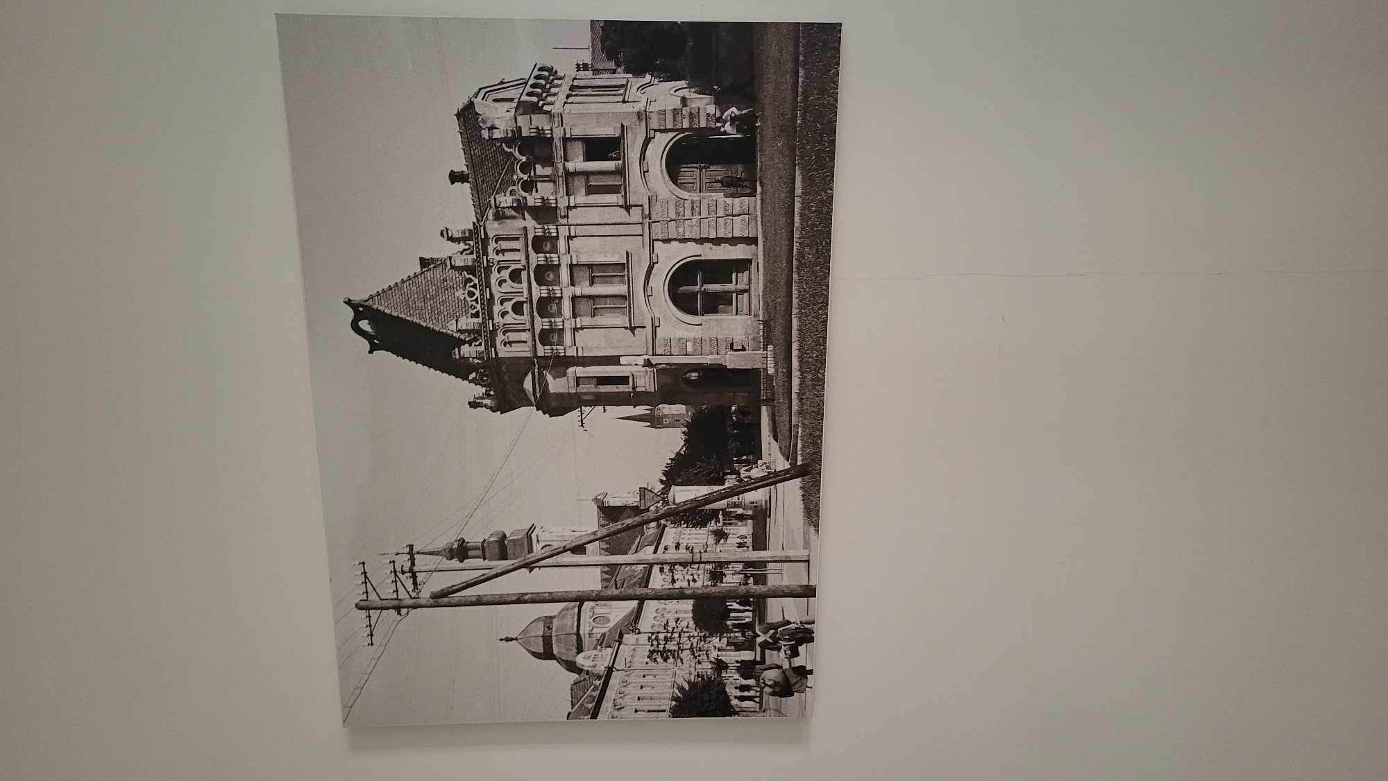The image is a black and white photograph mounted on a wall. It depicts a historic street scene in Prešov, Slovakia, likely taken in the early to mid-20th century. In the foreground, a wooden utility pole with insulators and wires stands prominently, bisecting the frame slightly to the left. To the left of the pole, a row of buildings lines a street, characterized by classical architecture, arched windows, and domes, suggesting institutional or public structures. Several people are visible walking on the sidewalk, some carrying parcels, indicating daily life and commerce. In the mid-ground, the prominent building on the right is a large, ornate structure with a steeply pitched roof, decorative stonework, and multiple windows, featuring arched entrances at street level. To the left of this building, a spire of a church or similar tower rises above the other structures. The overall environment appears to be an urban center with a mix of architectural styles typical of Central Europe.