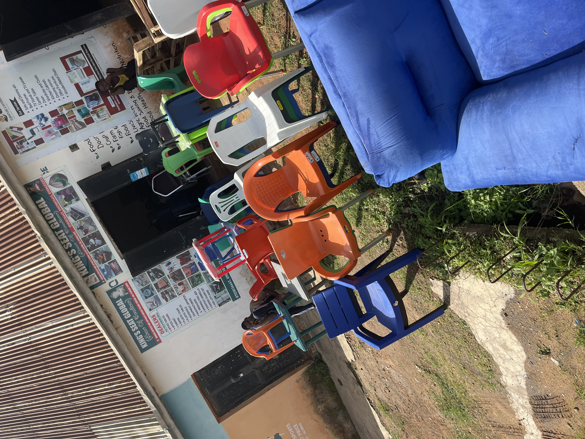 The image depicts the exterior of a business named "KING'S SEAT GLOBAL" located in Bukuru, Nigeria. The storefront features a large sign with the business name and various advertisements for furniture. In front of the store, an assortment of colorful plastic chairs, both for children and adults, are displayed on the ground. Some of these chairs are stacked, while others are arranged around small tables. A young boy is seated on one of the plastic chairs, looking towards the camera. Behind the chairs, a blue upholstered sofa is visible, suggesting a broader range of furniture offerings. The environment appears to be a dusty, outdoor area with some patches of grass and vegetation. The building itself has a corrugated metal roof, characteristic of many structures in the region. The overall scene conveys a busy marketplace atmosphere where furniture is sold outdoors.