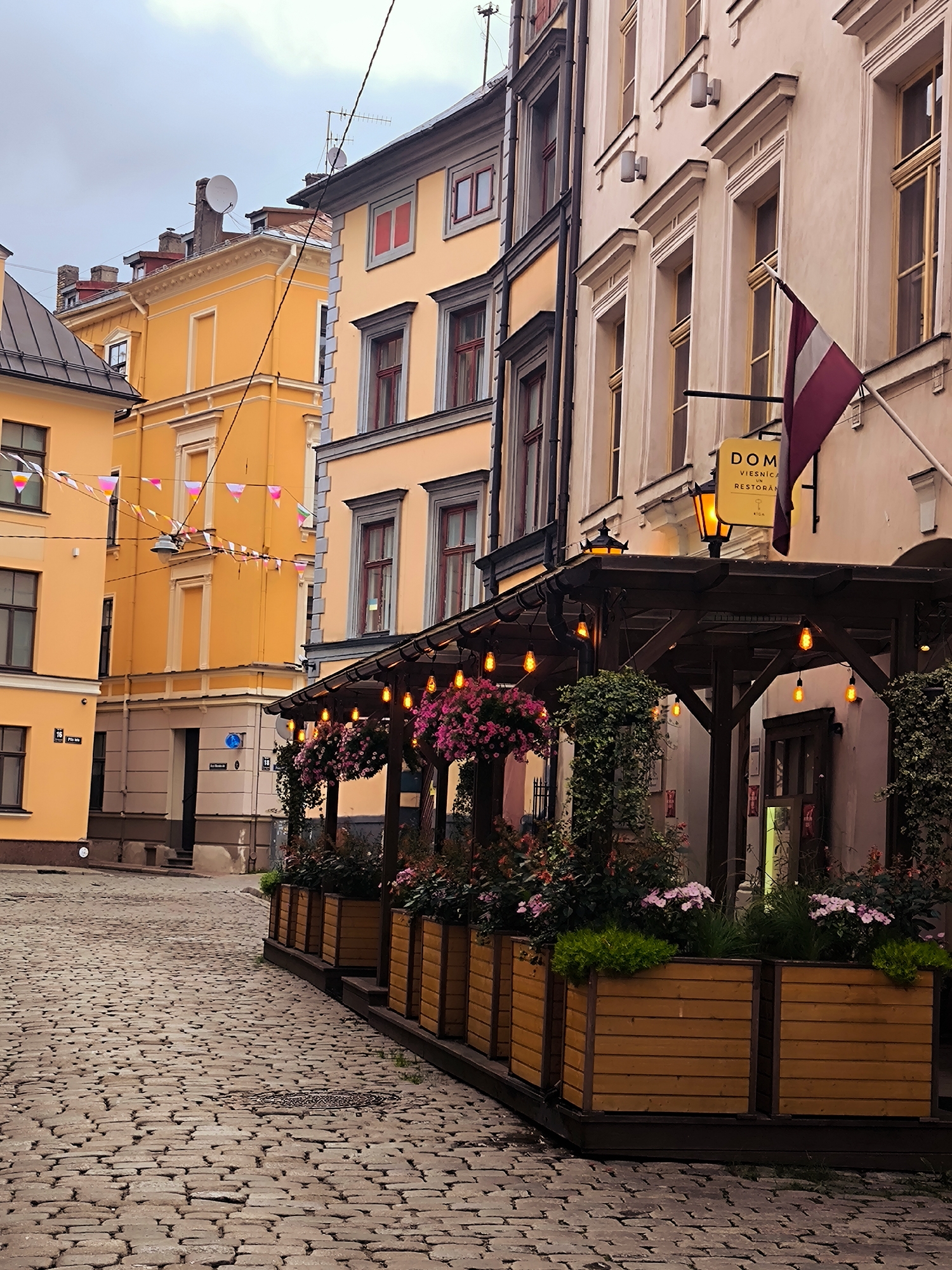 The image depicts a narrow cobblestone street in the Old Town of Riga, Latvia, during overcast daylight. On the right, a restaurant with an outdoor seating area is visible. The seating area is covered by a wooden canopy adorned with string lights and hanging baskets of pink flowers. Wooden planters filled with greenery and flowers line the front of the seating area. A sign for "Dom" restaurant is mounted on the building facade, alongside the Latvian flag. The buildings on both sides of the street are a mix of light yellow and cream colors, with traditional architectural details and windows. Bunting is strung across the street in the background. The cobblestone pavement is well-worn, suggesting a historic location.