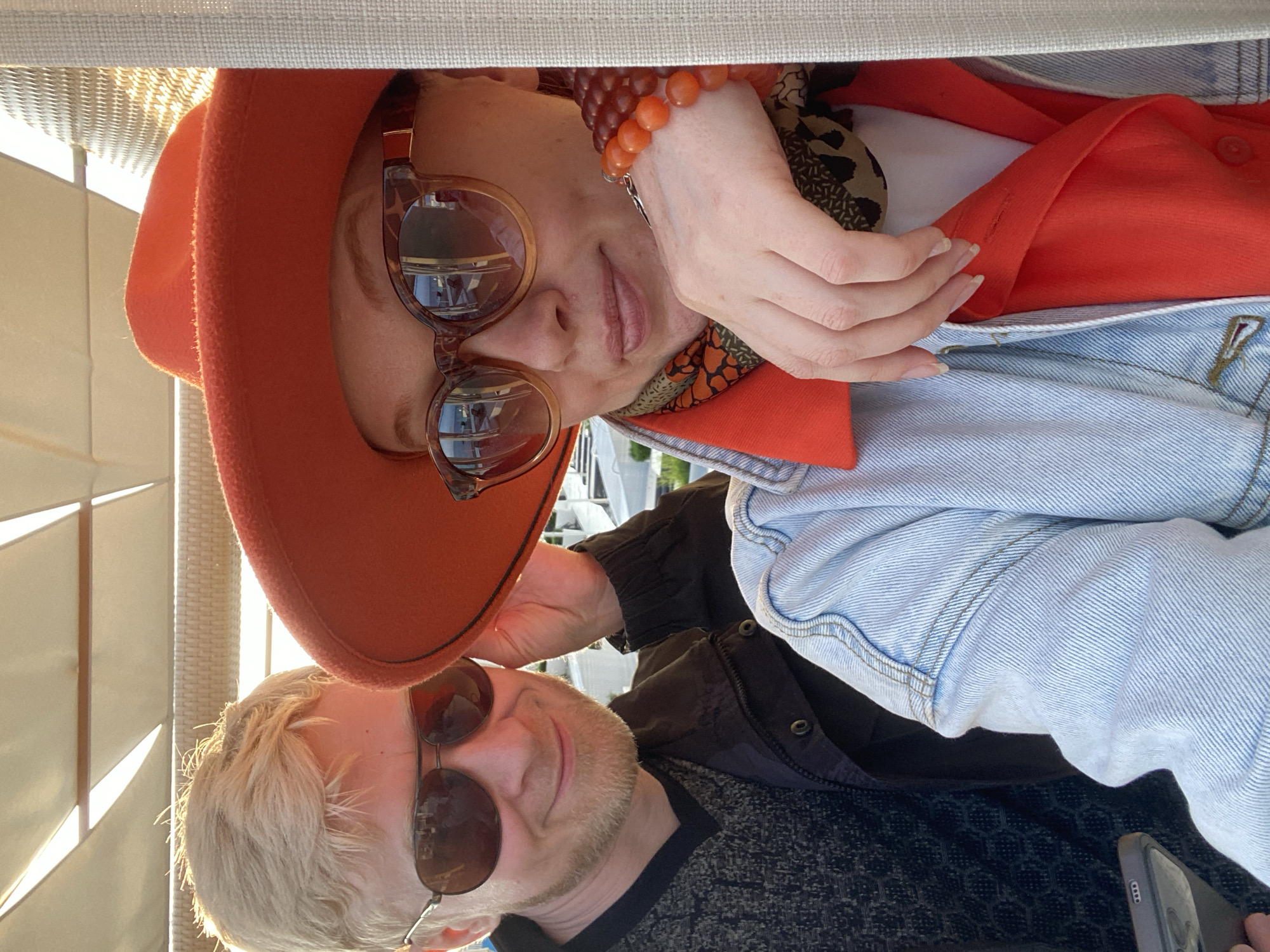 A photograph depicts two individuals, likely a couple, in an outdoor setting in Sochi, Russia, as suggested by the Russian Cyrillic letters "СОЧИ" faintly visible on a sign reflected in the woman's sunglasses. The woman is wearing an orange hat, patterned sunglasses, an orange top under a light blue denim jacket, and a beaded bracelet. The man has blonde hair, sunglasses, and is wearing a dark patterned shirt with a dark jacket. They are posing for a selfie, with the woman holding her hand close to her face. The background is composed of a lattice-like structure and a white covering, possibly part of an outdoor seating area.