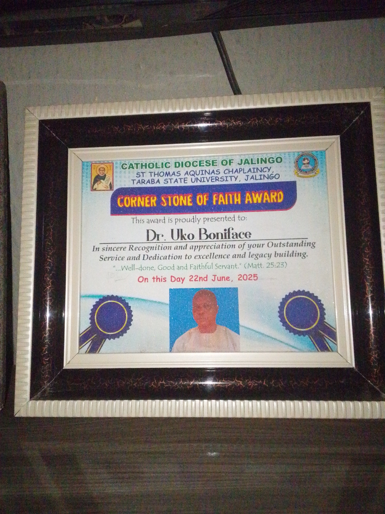 This is a photograph of a framed award certificate. The certificate is for a "Corner Stone of Faith Award" presented by the Catholic Diocese of Jalingo, St. Thomas Aquinas Chaplaincy, Taraba State University, Jalingo.
The award is presented to "Dr. Uko Boniface" in recognition of their "Outstanding Service and Dedication to excellence and legacy building." A Bible verse, "(Matt. 25:23)" is quoted on the certificate. The award is dated "22nd June, 2025."
The certificate features a small icon of a religious figure in the upper left corner and a circular emblem in the upper right corner. Below the recipient's name, there is a photograph of a man wearing traditional white attire and a cap. Decorative blue and yellow ribbons are featured at the bottom corners of the certificate.
The frame itself is brown and white with a patterned inlay. The photograph appears to be taken indoors, possibly in a home or office, with a dark wooden surface visible in the foreground. The lighting suggests it is daytime, and there are no prominent environmental cues. The overall tone is formal and celebratory, befitting an award ceremony.
