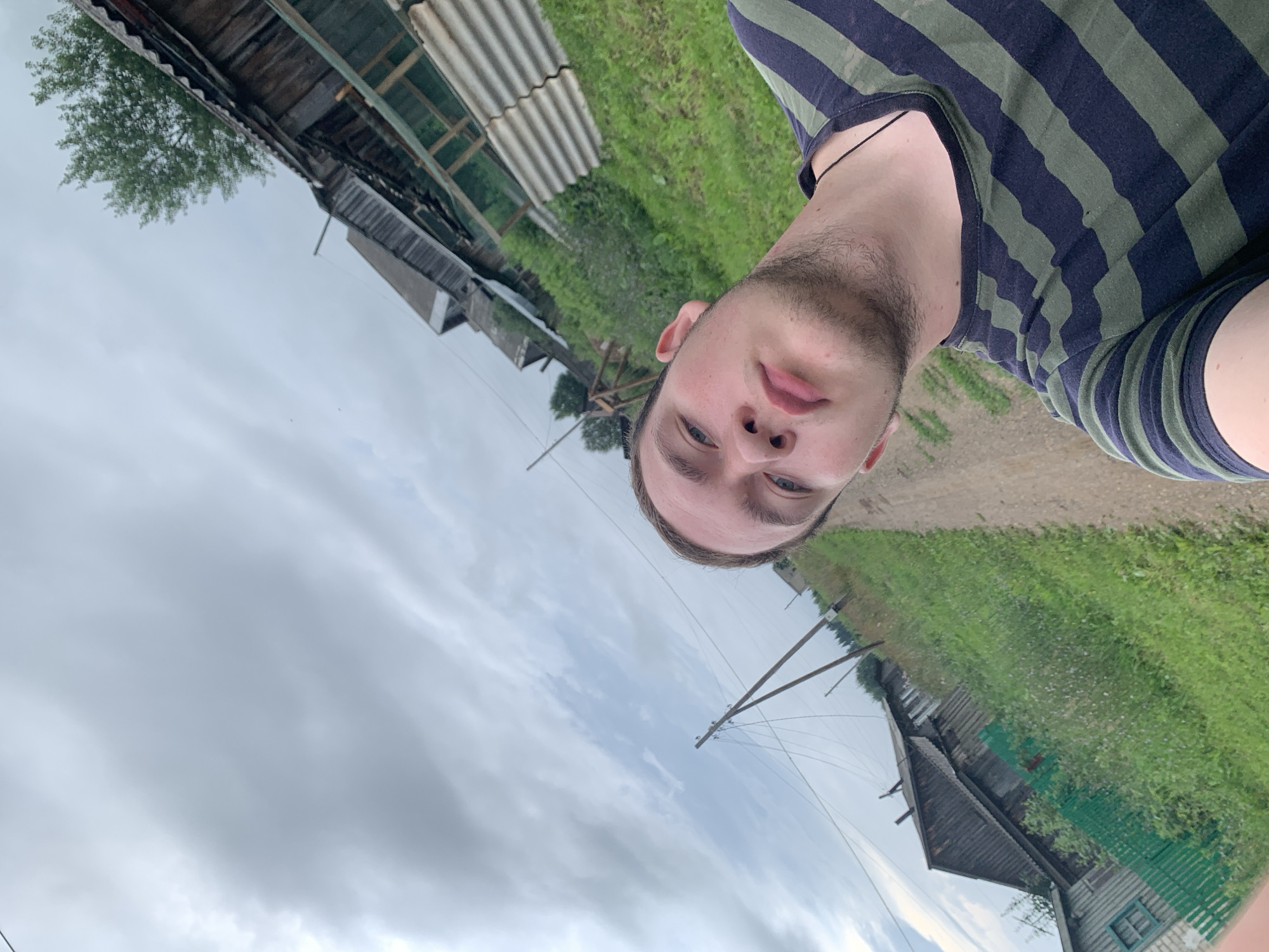 A selfie is taken by a young Caucasian male with short brown hair and blue eyes. He is wearing a dark green and navy striped t-shirt. He has a light stubble on his face and appears to be looking towards the camera. The background depicts a rural scene in Panteleevka, Russia. There is a dirt road running through the middle of the frame, with grassy areas on either side. Several wooden structures, likely houses and outbuildings, are visible in the distance. Electrical poles with wires are also present. The sky is cloudy and overcast.