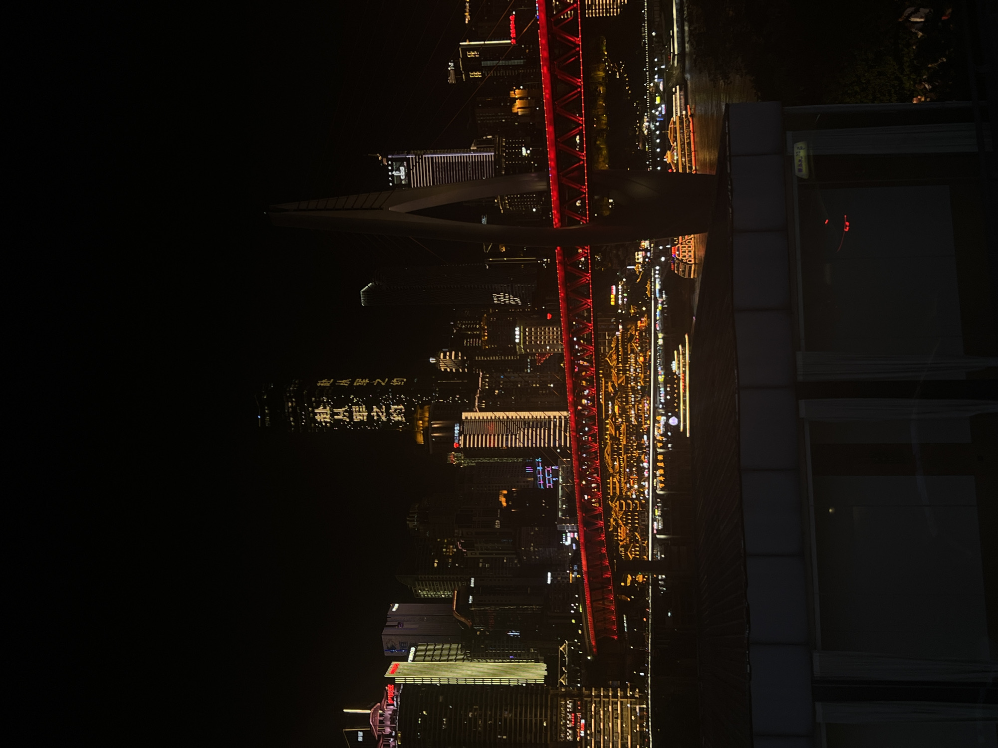 This is a nighttime photograph of a city skyline, possibly taken from a high vantage point. The image is oriented vertically, with the bottom of the frame showing the dark water of a river or bay, reflecting the illuminated cityscape. Across the water, a dense collection of modern buildings, many of them skyscrapers, are lit up with a variety of lights, including white, yellow, and red.
A prominent feature of the scene is a large, illuminated red bridge spanning across the water, with intricate latticework and bright red lights. The bridge appears to connect different parts of the city. In the foreground, there is a dark structure that might be a balcony or part of a building, with large windows that reflect the city lights and show some interior details.
The overall impression is one of a vibrant, bustling urban environment at night. The time of day is clearly night due to the darkness and the prominent illumination of the city lights. The weather appears clear, allowing for good visibility of the cityscape and its reflections.
Visible text in Chinese characters can be seen on some of the buildings and possibly on signage, though the text is not entirely legible in the provided image. For example, there are large characters written vertically on a building in the middle left of the image, appearing to be illuminated by yellow lights. Another sign with yellow text is visible on the foreground structure on the right. The location context suggests the scene is in Jiangbeicheng Subdistrict, China.
