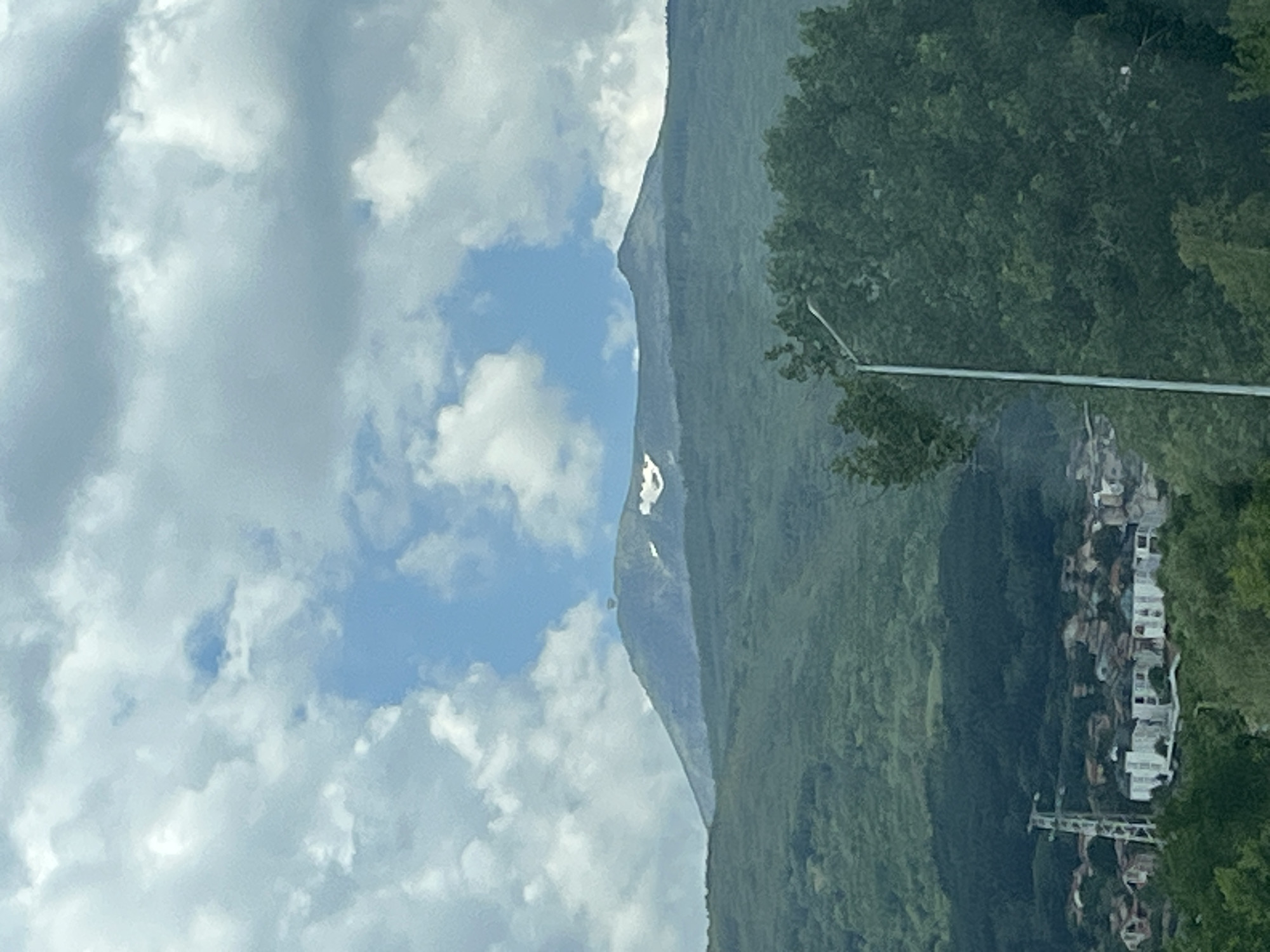 The media file displays a scenic view of a mountain range partially obscured by clouds. The mountain is covered in lush green vegetation, suggesting a tropical or temperate climate. A narrow strip of white, possibly a waterfall or a snow patch, is visible on the mountain's face.
Below the mountain, on the right side of the frame, a cluster of buildings with white facades is nestled into the hillside. The architecture appears to be contemporary with multiple stories. A white pole, possibly a streetlight or part of a utility line, extends horizontally across the middle right of the image, with some rigging or structure visible below it.
The sky is partly cloudy, with large, fluffy white clouds interspersed with patches of blue sky, indicating fair weather. The lighting suggests it is daytime, though the specific time is not discernible. The overall mood is serene and picturesque, capturing the grandeur of nature and the human element in harmony with it. No people are clearly visible in the image. There is no discernible text in the media file.