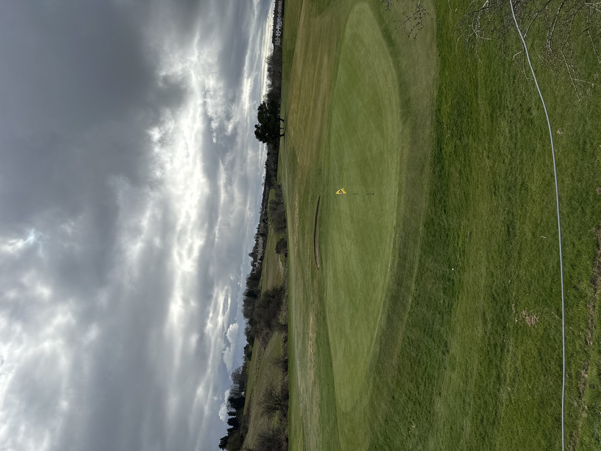 The image shows a golf course in Dunfermline, United Kingdom. The foreground consists of a large, green grassy area with a yellow flag marking a hole. The background shows trees, a grassy hill, and what appears to be buildings in the distance. The sky is overcast with grey clouds. A grey electrical cable runs from the upper right corner into the image. There is no evidence of people.
