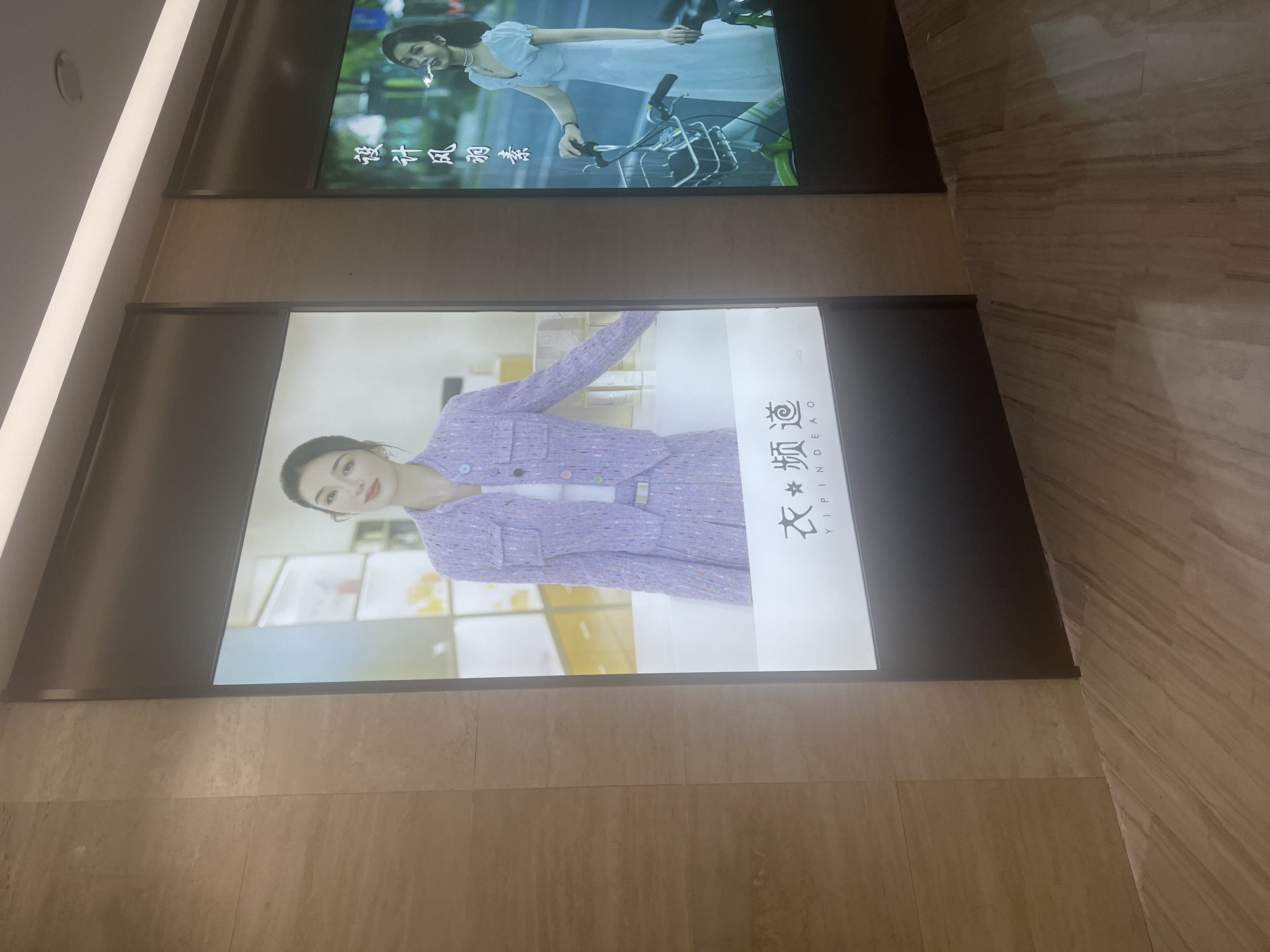Two large digital billboards are displayed in a modern, indoor commercial space with marble-tiled walls and flooring. The billboard on the left features a woman wearing a lavender tweed suit and a white top, posing with a soft smile. The billboard on the right shows a woman in a white dress, holding onto a bicycle with a basket, and appearing to smoke a cigarette, with Chinese text that translates to "Design, Style, Purity" above her. The context suggests these billboards are likely part of a retail establishment or shopping mall in Liandu District, China, promoting fashion or lifestyle brands.