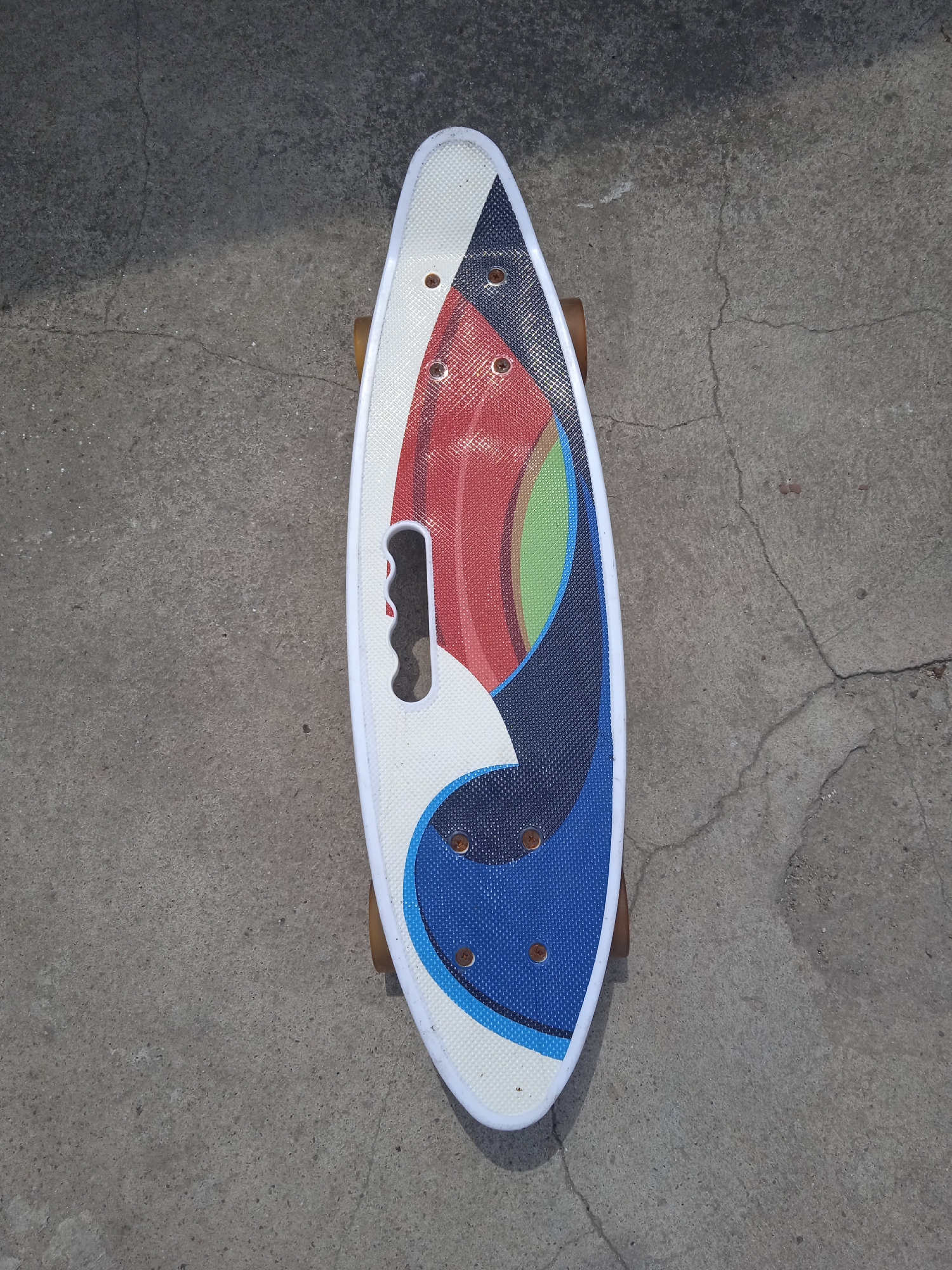 A top-down, full shot shows a white and multi-colored skateboard lying flat on a grey concrete surface. The skateboard is a penny board, a short and narrow skateboard, with rounded ends. It has a white grip tape top with a cut-out handle in the middle. The graphic on the skateboard consists of abstract shapes in red, blue, green, and white. The wheels, one brown one visible, are attached to the underside. The concrete surface is cracked and weathered, with a shadow cast diagonally across the scene from the upper left to the lower right.