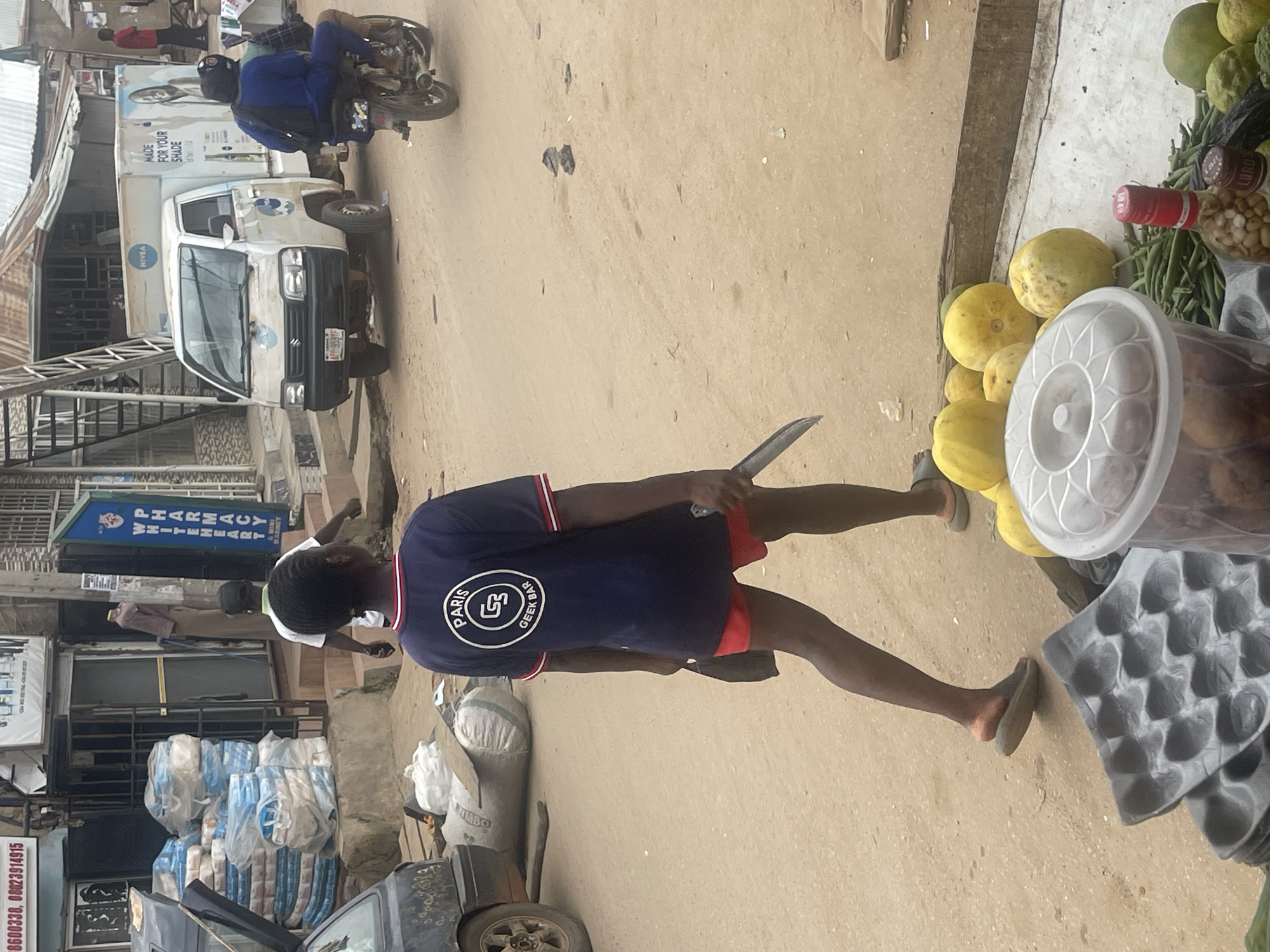 A person wearing a dark blue shirt with "PARIS GEEK BAR" logo and red shorts walks to the left on a dirt road. The person is holding a large knife in their right hand and a dark bag in their left hand. The person is wearing dark sandals. In the background, a white truck with "NIVEA" branding is parked. Several bags of products are stacked in front of a store. A motorcycle with a passenger is seen on the right side of the frame. Fruits and vegetables are laid out on the ground in the foreground. The scene appears to be a street market in Abuja, Nigeria.