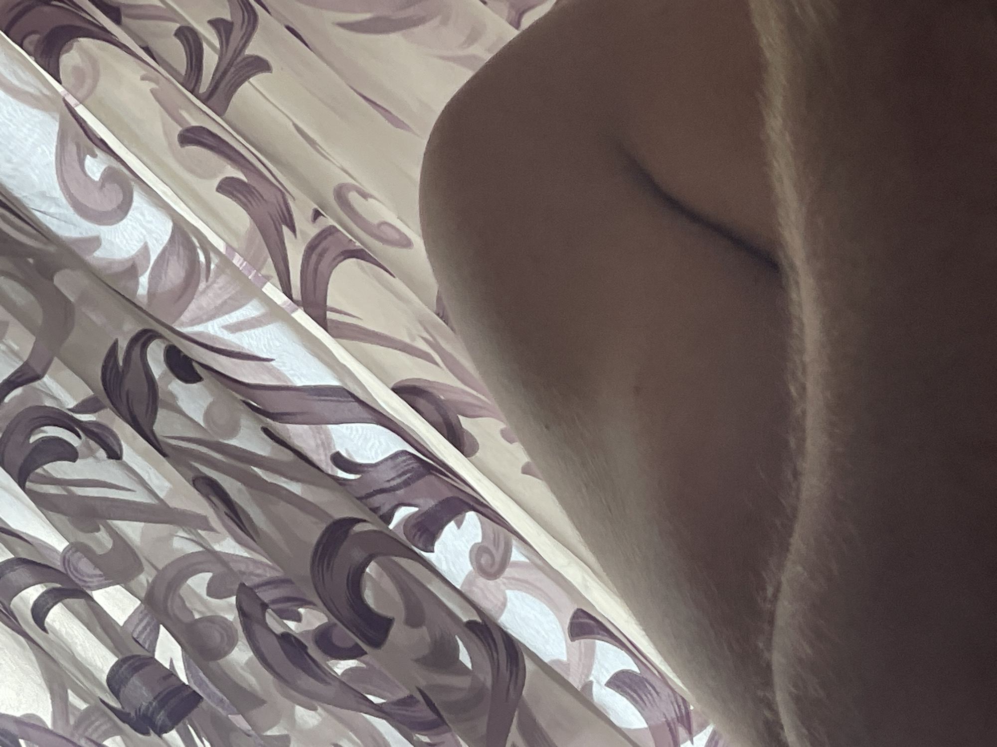 The image shows a close-up view of a person's knee and thigh, with a patterned curtain in the background. The curtain has a swirling, floral design in shades of purple and white. The skin is lightly tanned and shows some texture. The lighting suggests the image was taken indoors, possibly near a window. There is no visible information within the image to indicate its location in Varna, Bulgaria.